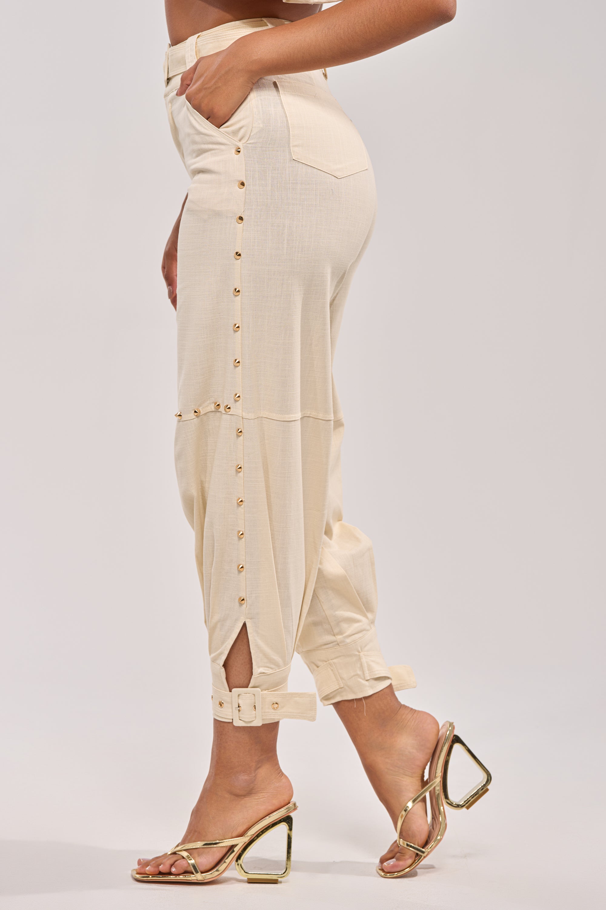 A person models the MAEVE LINEN STUDDED PANT, featuring a high waist, cream hue, belt, side buttons, ankle buckle strap, and metallic gold heels. Side view shown from torso to feet. 3