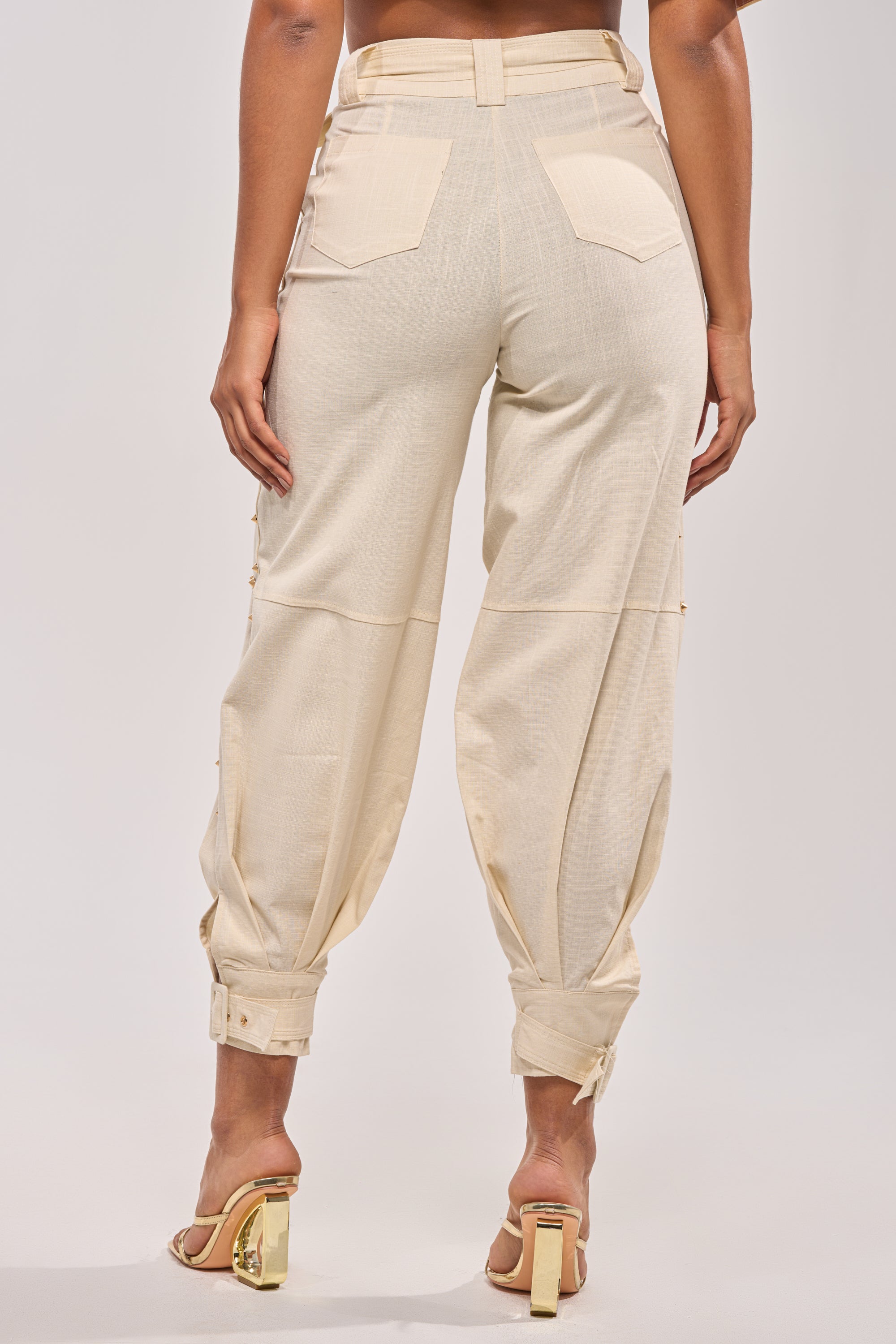 A person stands facing away, wearing MAEVE LINEN STUDDED PANT—high-waisted beige joggers with pockets and adjustable cuffs—paired with transparent-strap heeled sandals. 4