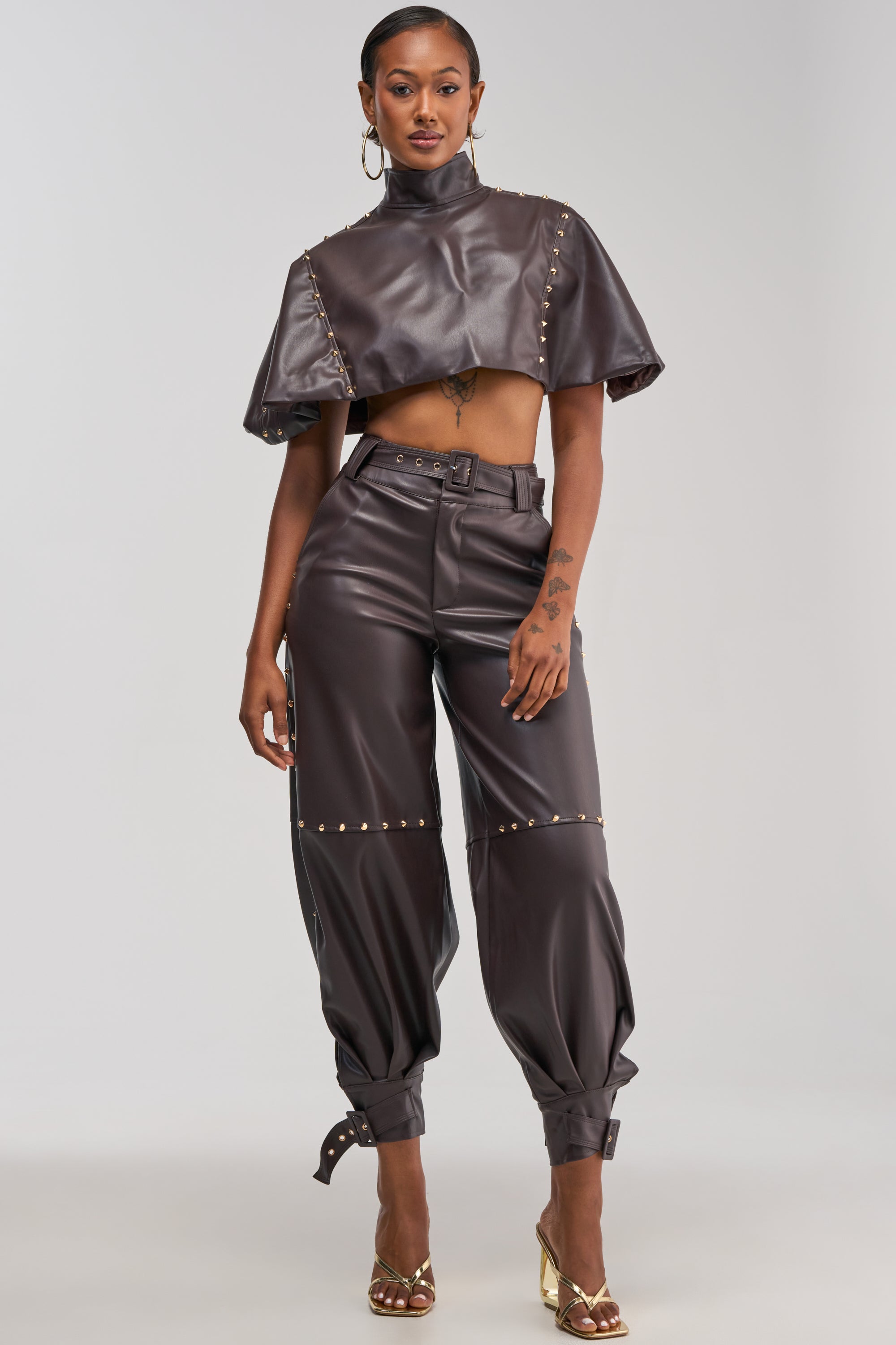 A woman stands confidently against a plain light background wearing gold hoop earrings, gold heels, a studded faux leather crop top with gold studs, and the STUDDED FAUX LEATHER BELTED PANT IN DARK CHOCOLATE.
