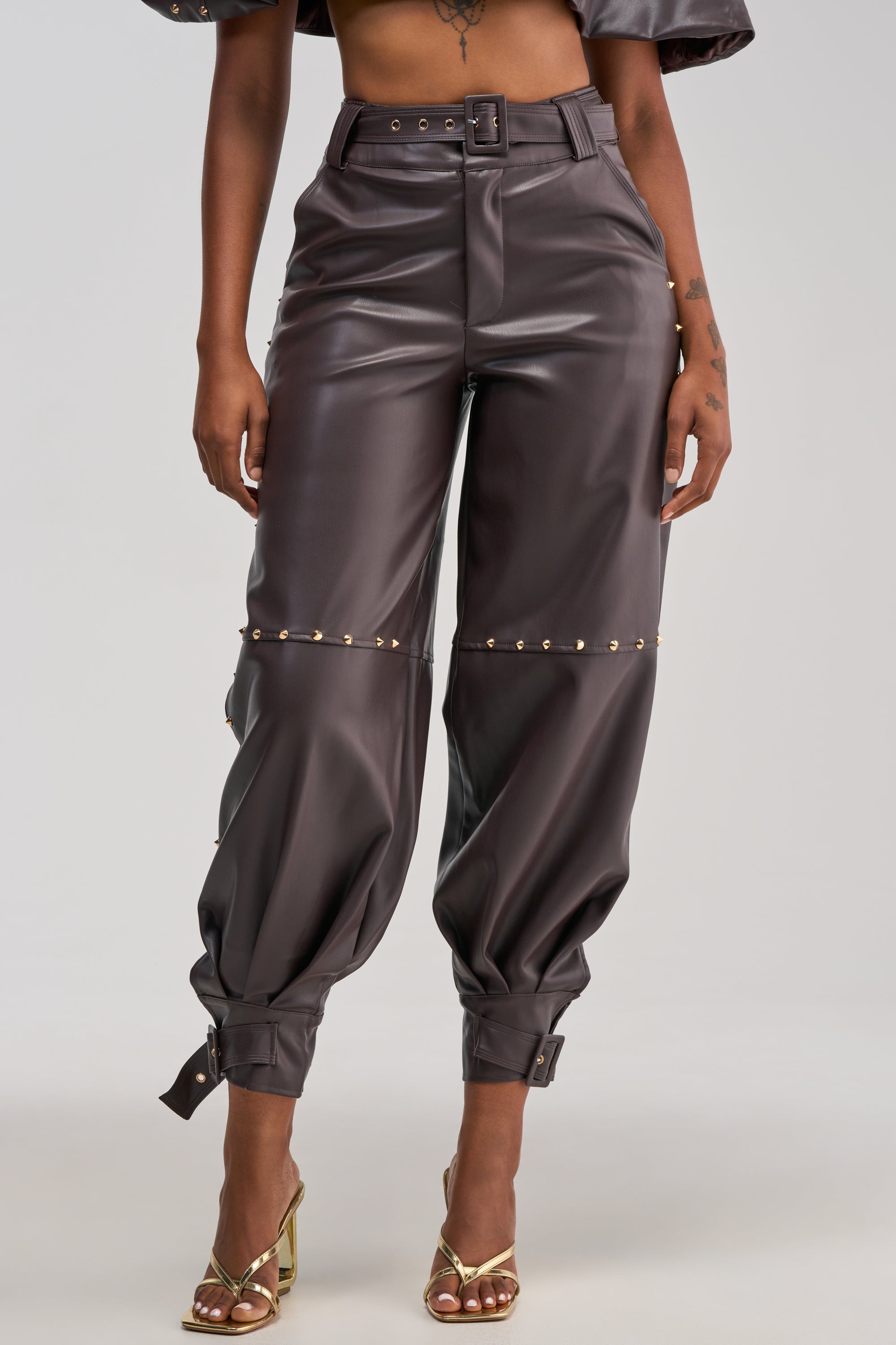 A woman models the STUDDED FAUX LEATHER BELTED PANT IN DARK CHOCOLATE—high-waisted, brown faux leather pants with a glossy finish, gold stud details, and buckle accents at the ankles—paired with gold heeled sandals. The image is cropped from shoulders to feet.