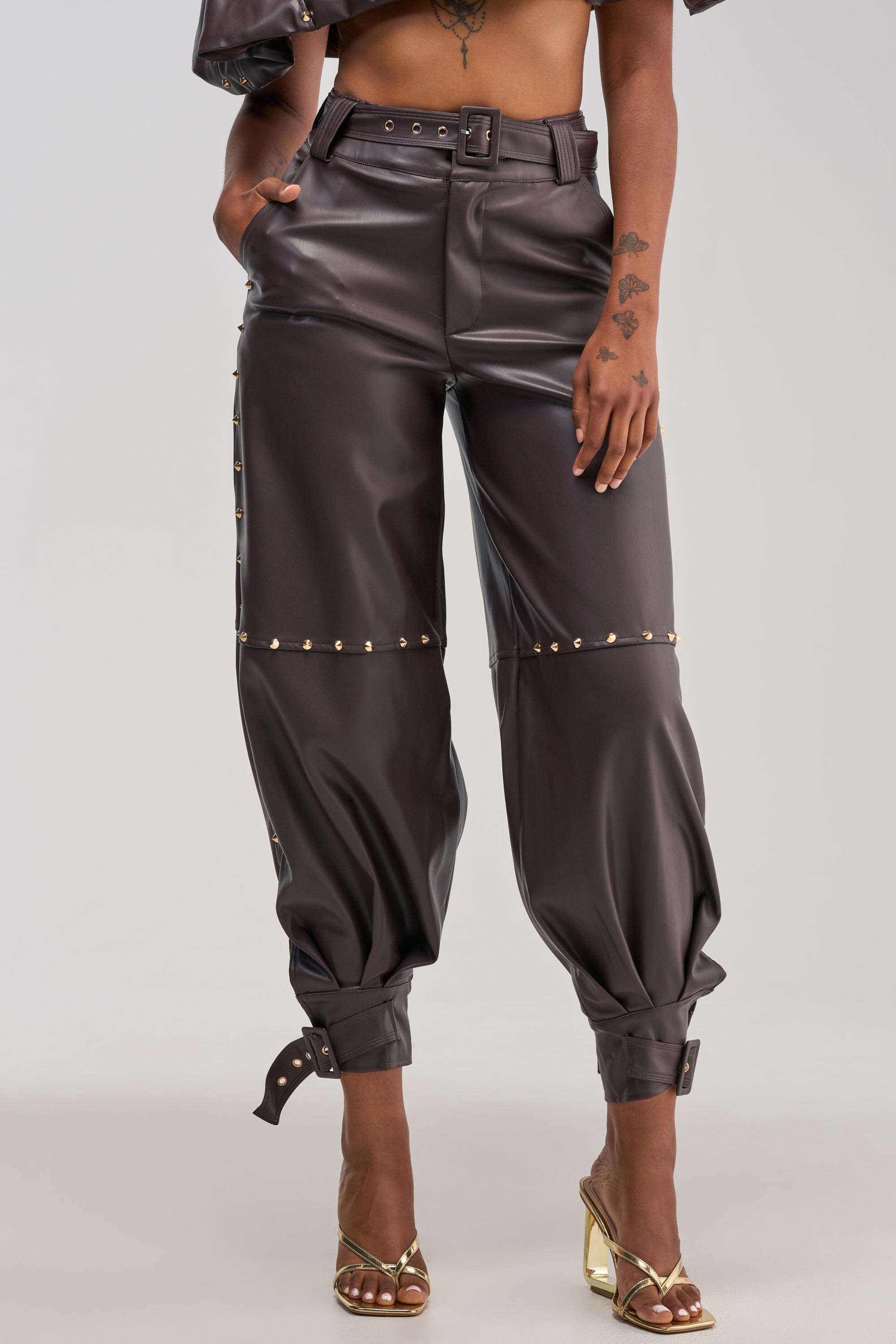 A person wearing the STUDDED FAUX LEATHER BELTED PANT IN DARK CHOCOLATE—high-waisted faux leather pants featuring gold studs and rolled cuffs—paired with gold open-toe heels.