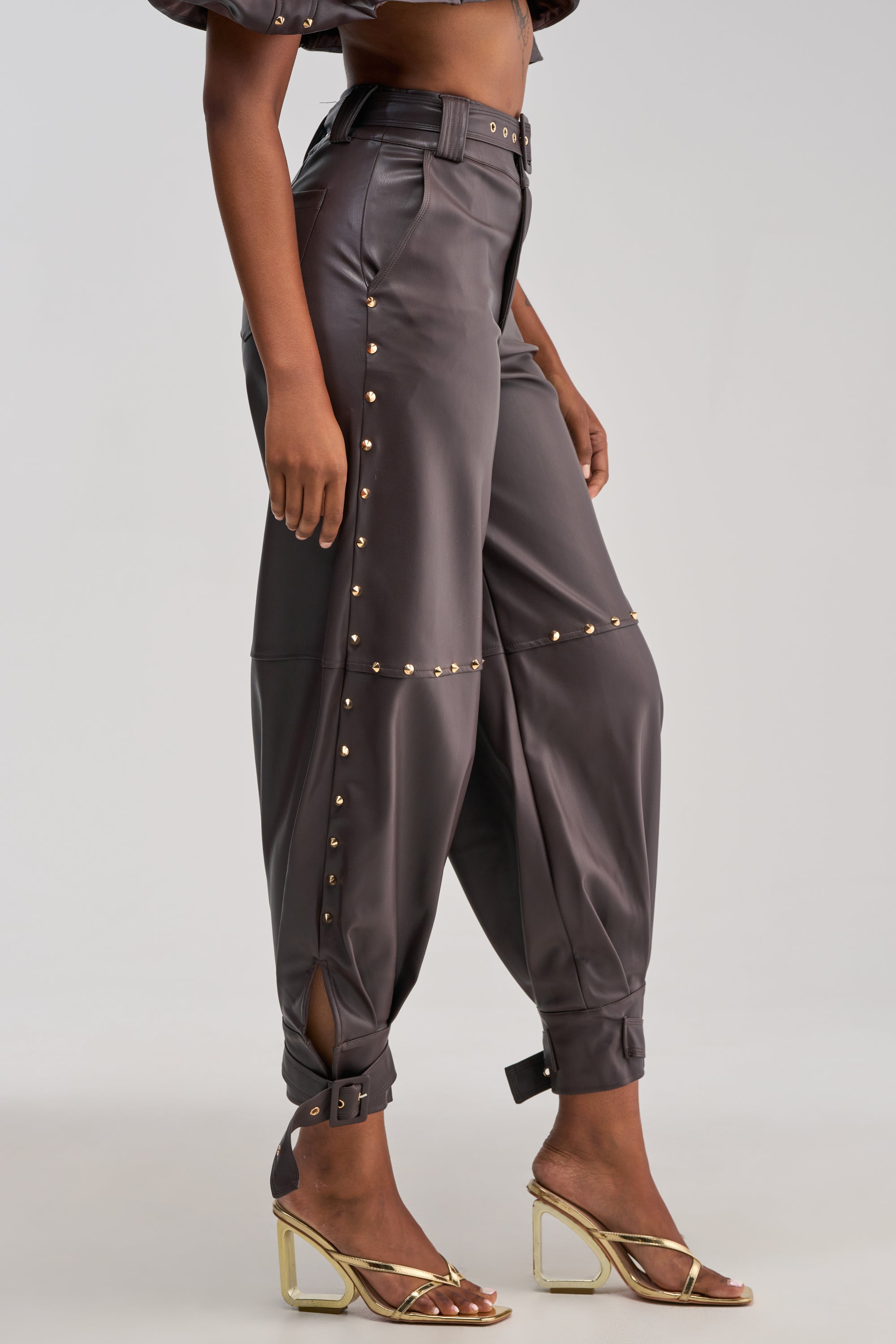 A woman models the STUDDED FAUX LEATHER BELTED PANT IN DARK CHOCOLATE—high-waisted pants detailed with gold studs and ankle straps—paired with gold heeled sandals. The image captures her from shoulders to feet against a plain background.
