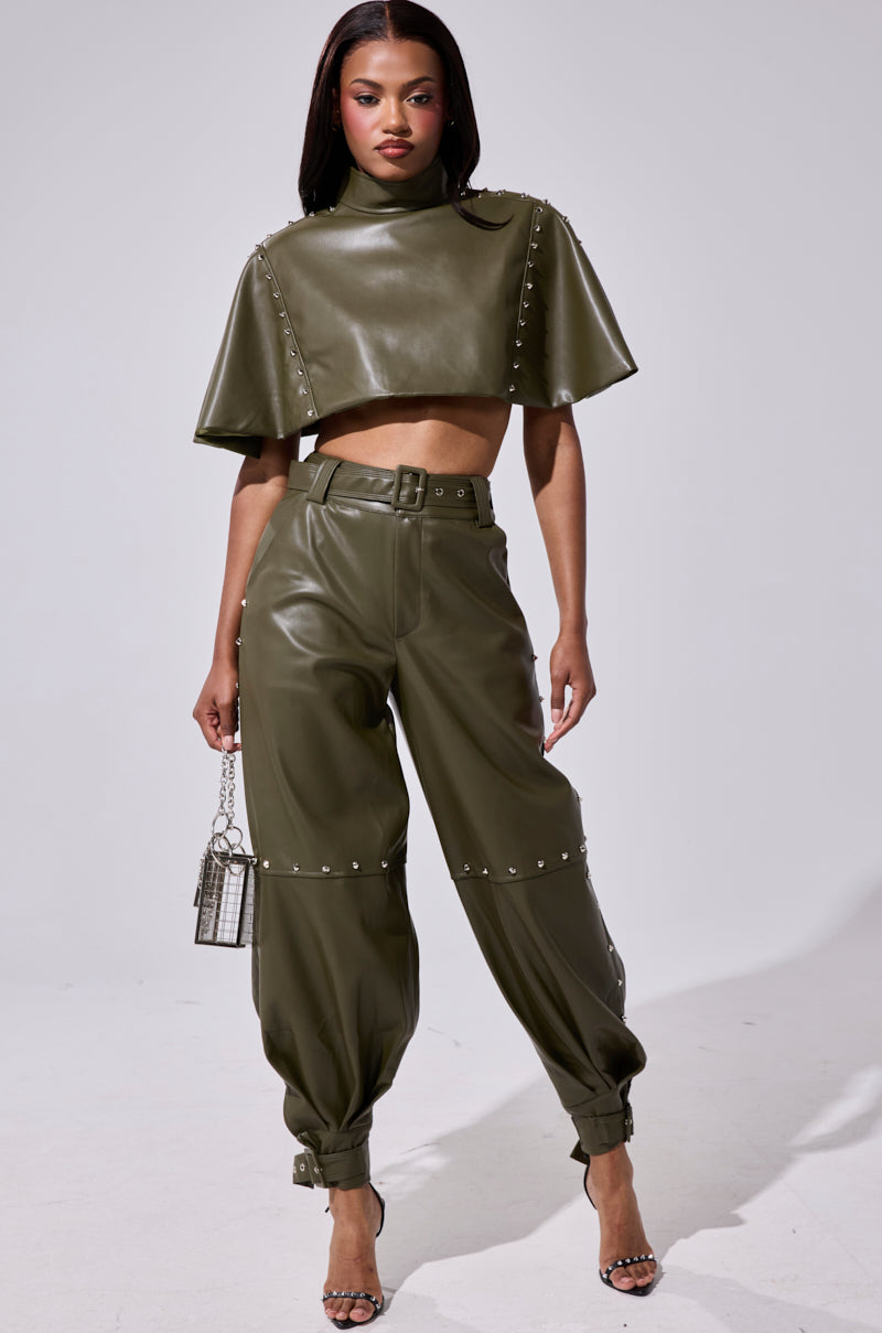 A woman exudes confidence in olive green STUDDED FAUX LEATHER BELTED PANT with a matching cropped, high-neck top, paired with a silver handbag and strappy heels.