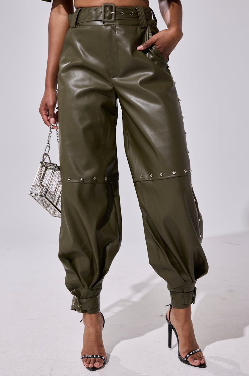 A person wearing STUDDED FAUX LEATHER BELTED PANT in olive green with buckle and snap details, paired with a silver handbag and black high-heeled sandals.