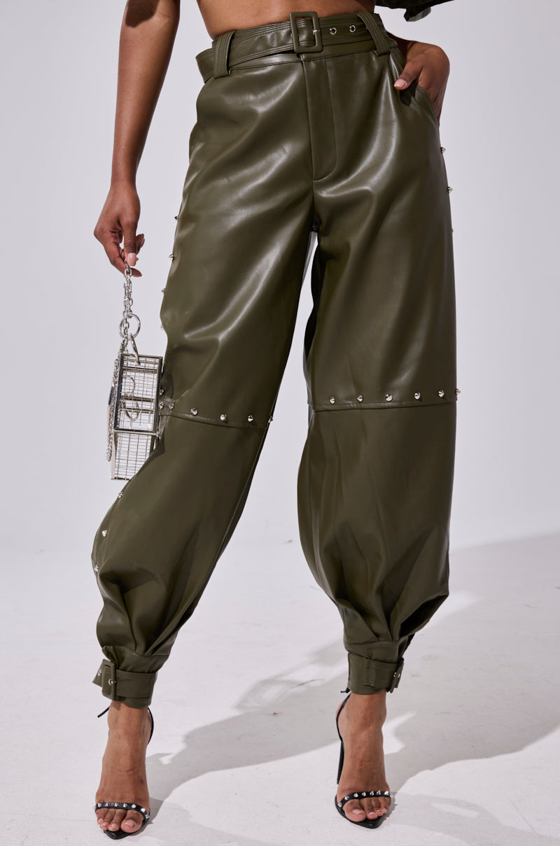 Wearing the STUDDED FAUX LEATHER BELTED PANT in high-waisted olive green with silver studs, ankle buckles, and a belted waist, paired with a metallic handbag and open-toe heels.