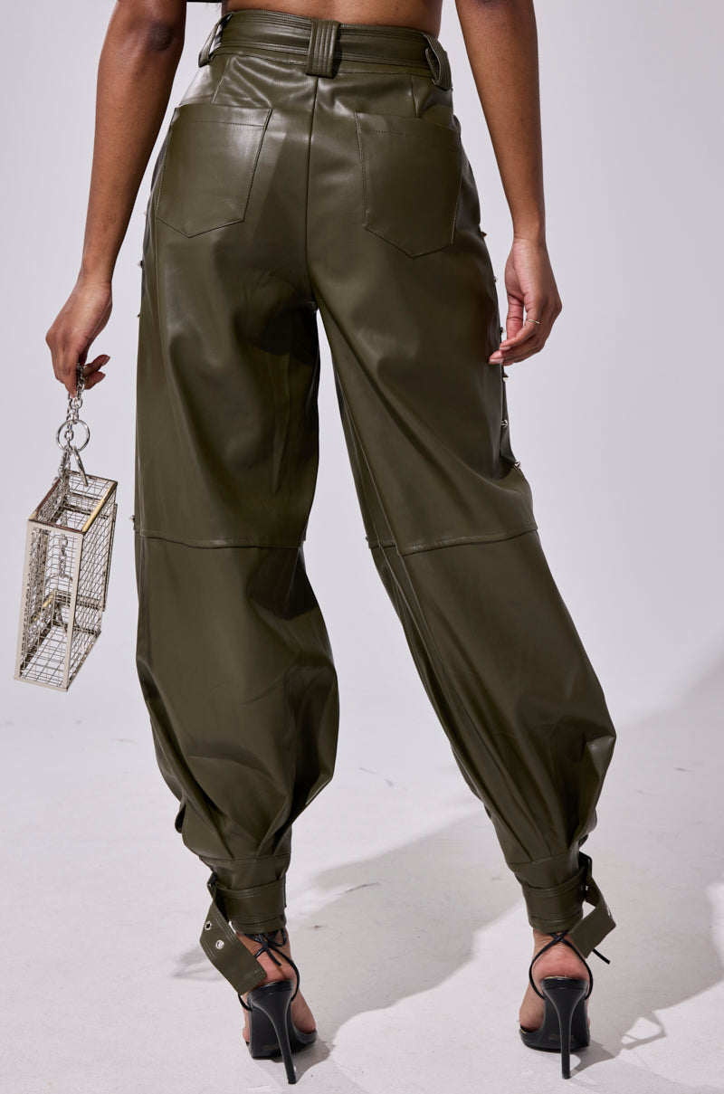 A person faces away, wearing high-waisted olive green STUDDED FAUX LEATHER BELTED PANT and black strappy heels, holding a small metallic geometric handbag in their left hand against a plain light background.