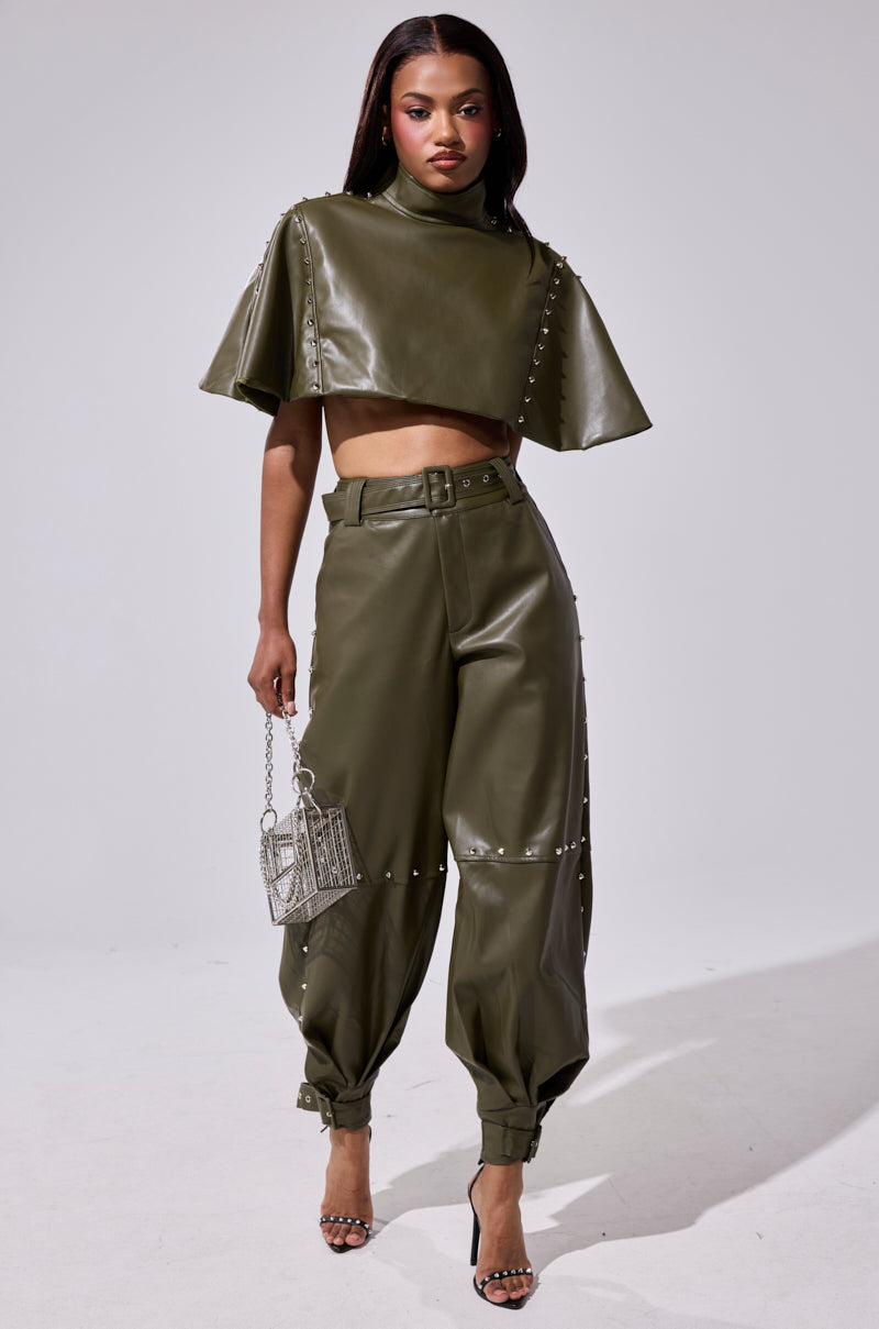 A woman stands confidently in olive green STUDDED FAUX LEATHER BELTED PANTS, paired with a matching crop top, strappy heels, and a metallic handbag. Silver studs add edgy detail to the bold two-piece ensemble.