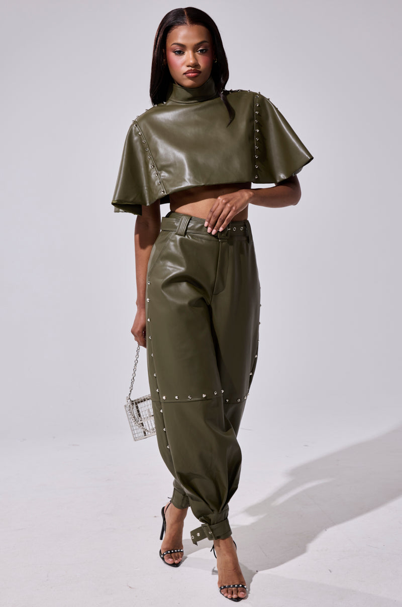 A woman wears the STUDDED FAUX LEATHER BELTED PANT in olive green with a cropped top, silver stud details, a small metallic purse, and high-heeled sandals.