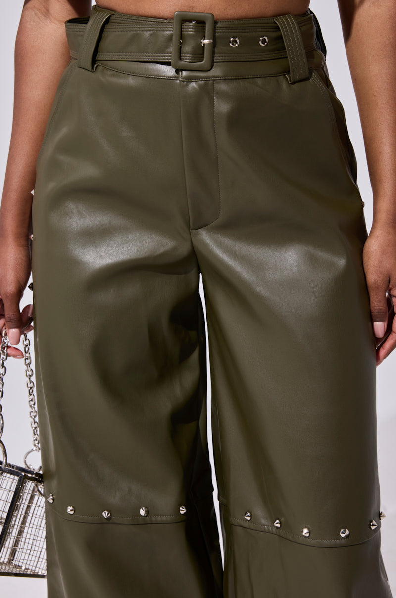Close-up of a person wearing the STUDDED FAUX LEATHER BELTED PANT in olive green with knee studs, paired with a small metallic chain-strap purse.