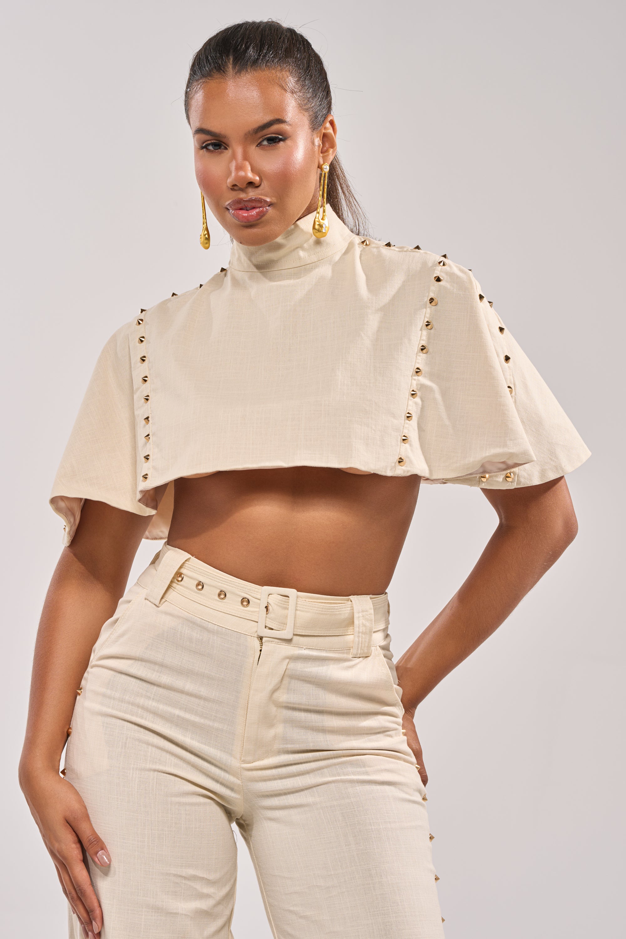 A woman models the MAEVE LINEN STUDDED BLOUSE—a cream, gold-studded cape top—paired with high-waisted pants and statement gold earrings. Her hair is pulled back, making it perfect for effortless summer style. 0