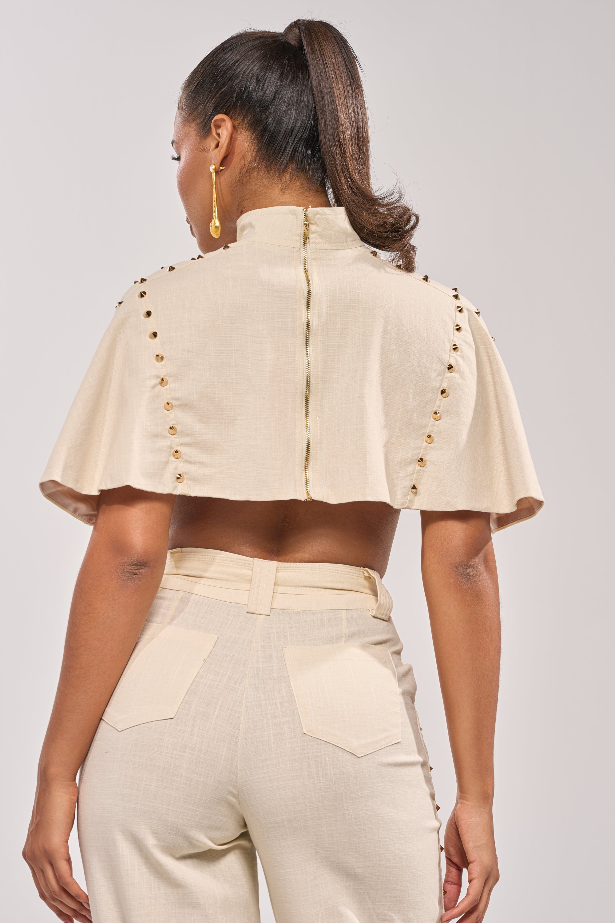 A woman is seen from behind in the MAEVE LINEN STUDDED BLOUSE, a light beige top with short cape sleeves and gold shoulder studs, paired with matching high-waisted pants. She styles her hair in a high ponytail and wears a gold earring—ideal for chic summer looks. 3