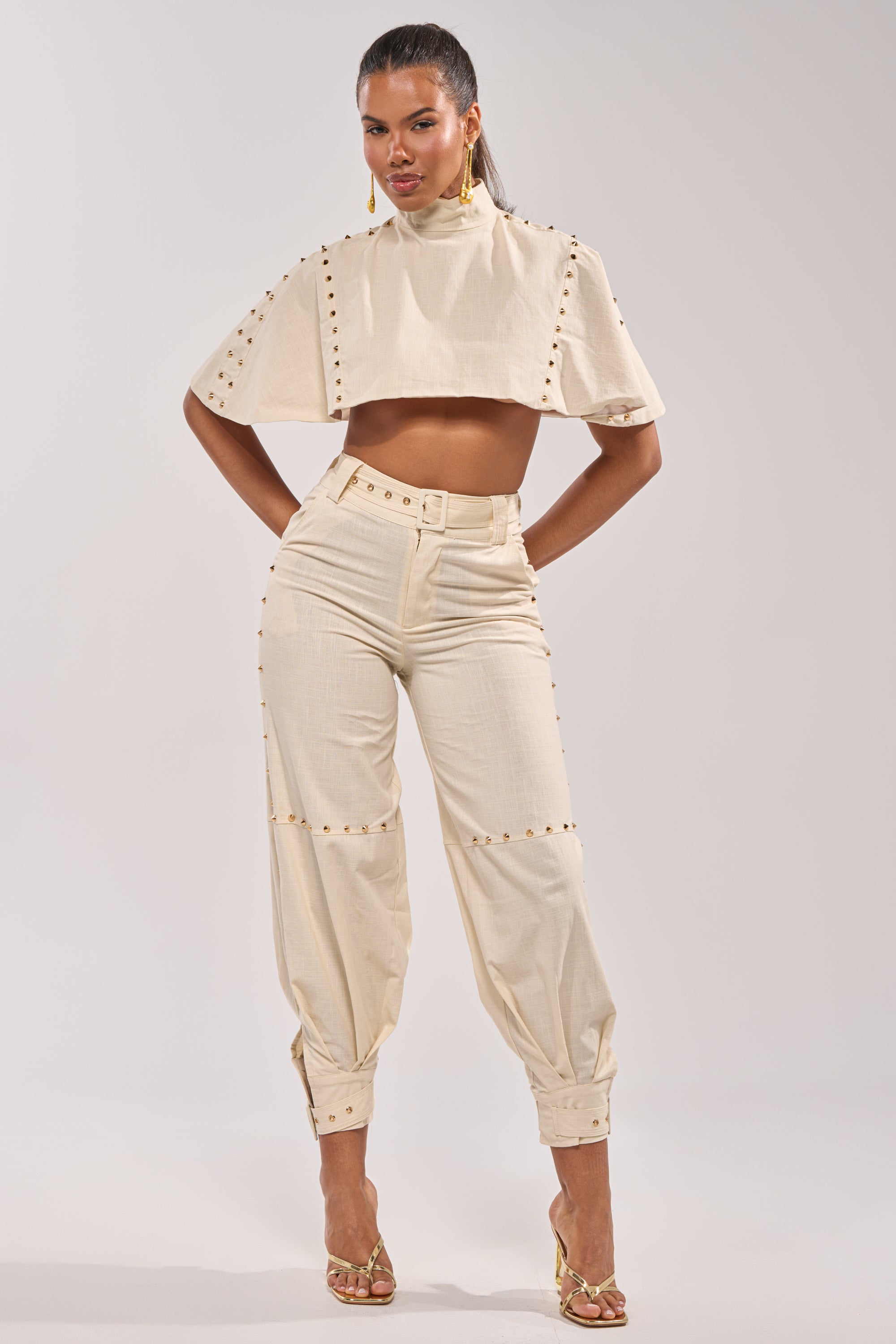 A woman stands confidently in the MAEVE LINEN STUDDED BLOUSE, paired with matching high-waisted pants, gold earrings, and strappy gold heels—an ideal summer look against a plain background. 4