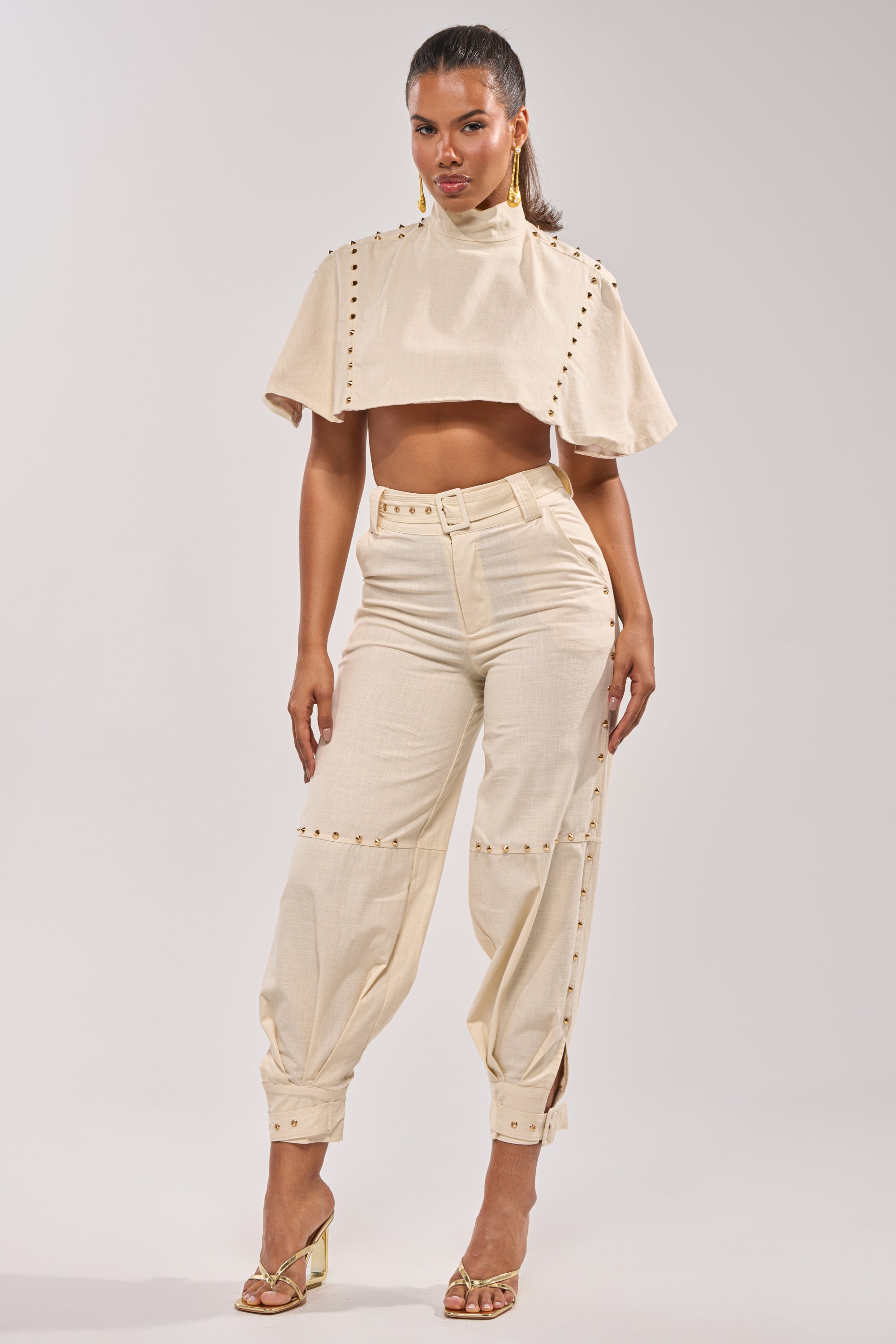 A woman models the MAEVE LINEN STUDDED BLOUSE in beige, paired with high-waisted, wide-leg pants. She accessorizes her summer look with gold earrings and heeled sandals, standing against a plain background. 5