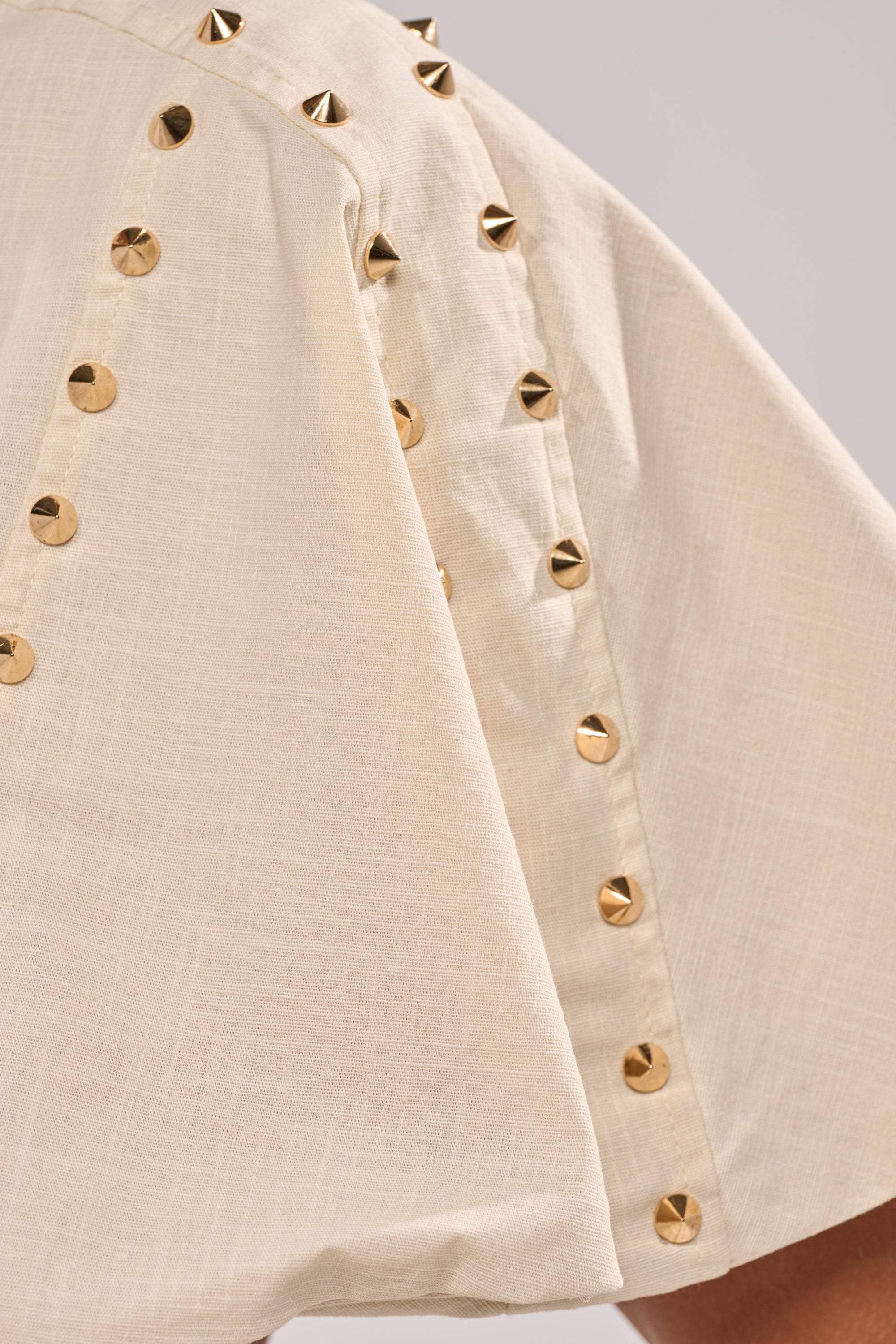 Close-up of the MAEVE LINEN STUDDED BLOUSE: a cream-colored linen cape featuring gold metal studs along the seams for a striking pattern—ideal as a statement top for easy summer style. 6