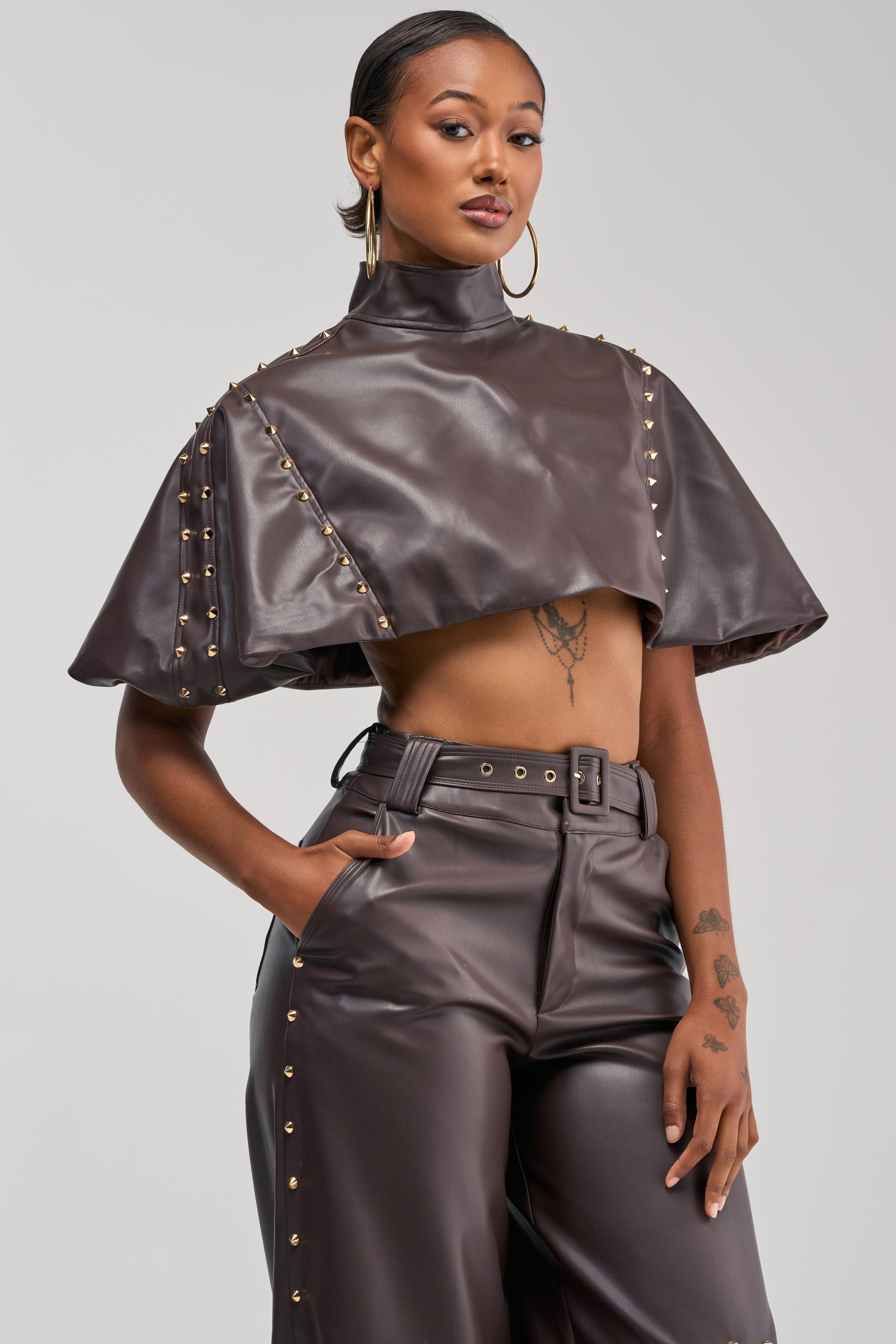 Wearing the Studded Faux Leather Cape in Dark Chocolate, a person poses against a plain background with one hand in their pocket. They style the look with matching high-waisted pants, gold hoop earrings, and show visible tattoos. The cape features gold-tone studs and a rich brown faux leather finish.