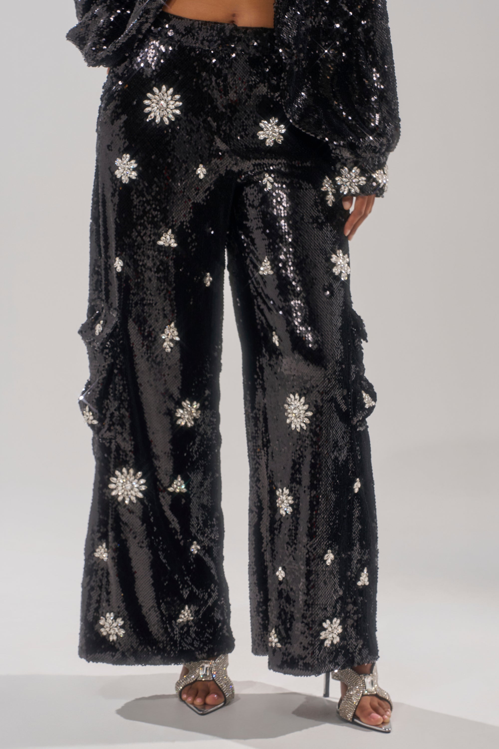 A cropped image shows a person’s lower body wearing DAZZLING HOLIDAY SEQUIN WIDE LEG CARGO PANT with black sequins and white floral accents, paired with matching open-toe high heels. 1