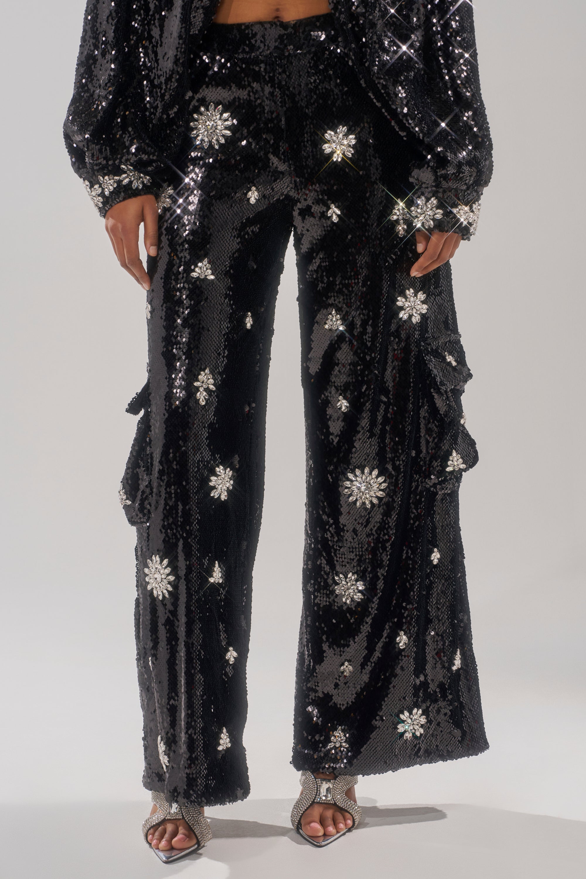 A person wearing DAZZLING HOLIDAY SEQUIN WIDE LEG CARGO PANT with silver starburst patterns, paired with matching high-heeled sandals and a sequined jacket. Image is cropped from the shoulders down. 2