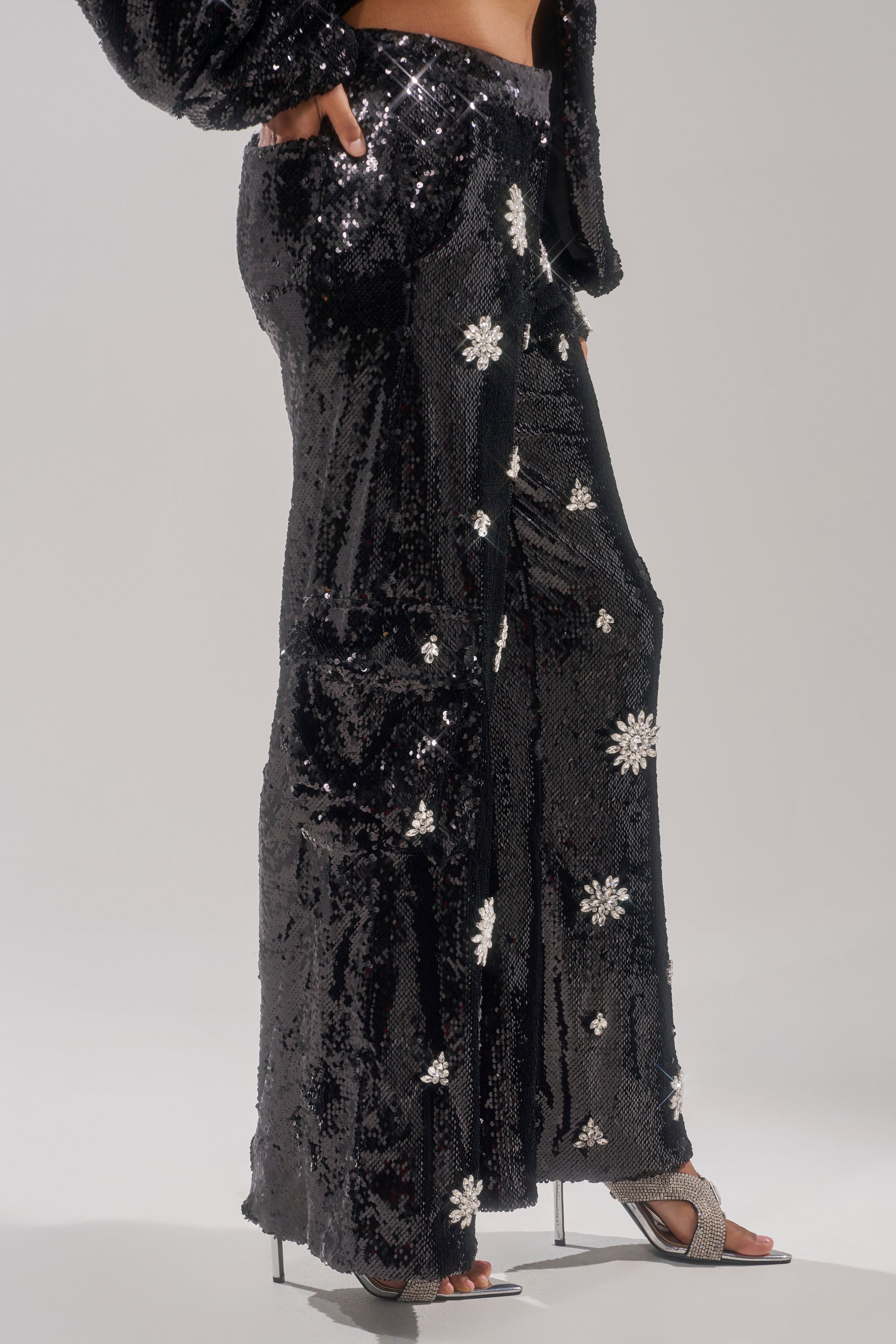 A person models the DAZZLING HOLIDAY SEQUIN WIDE LEG CARGO PANT, featuring white floral embellishments, styled with matching high-heeled sandals and striking a pose against a neutral background. 3