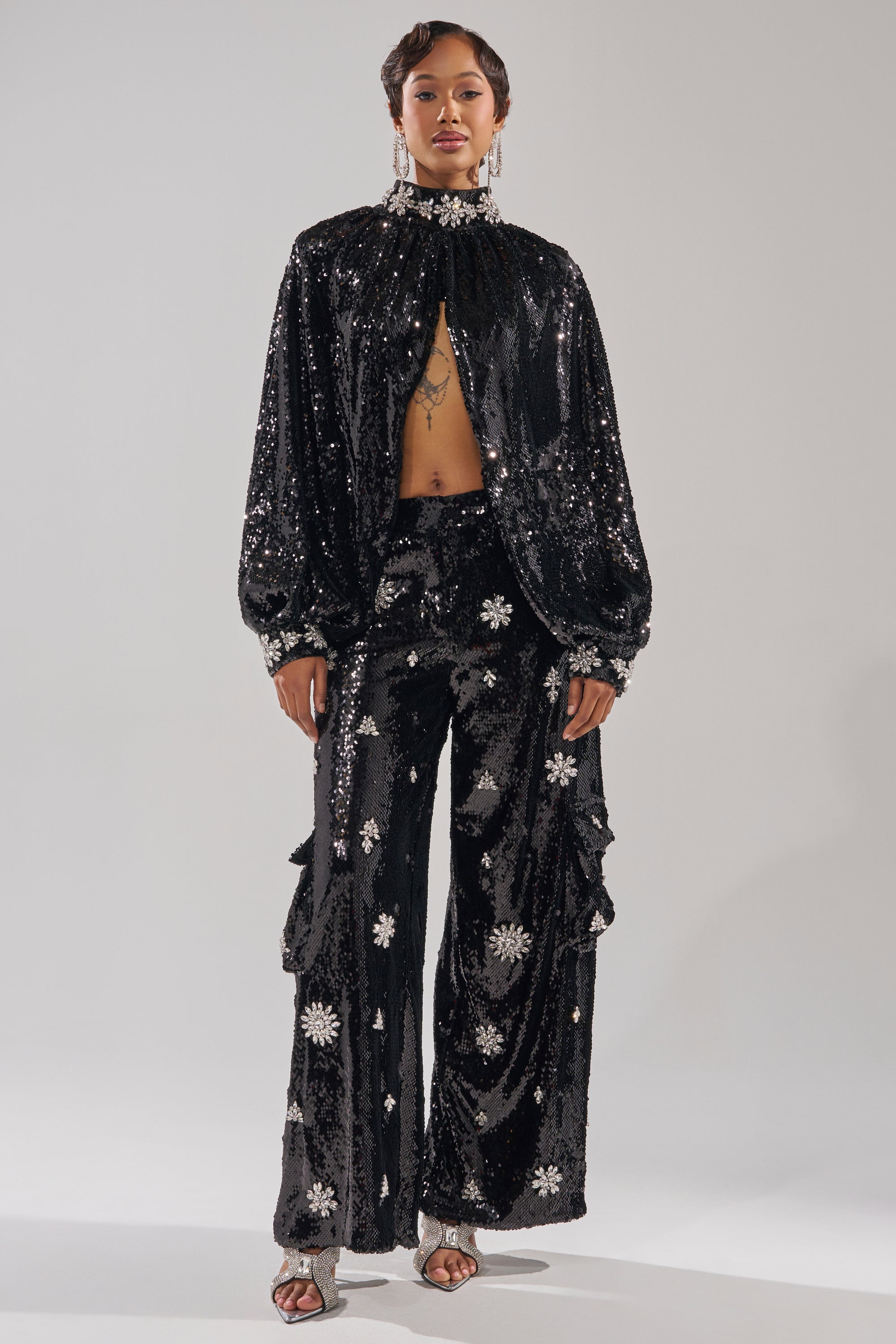 A person wears the DAZZLING HOLIDAY SEQUIN WIDE LEG CARGO PANT—a black, sequined wide-leg pant with silver starburst designs—paired with a high-neck, long-sleeve top, silver open-toe heels, and sparkly earrings on a plain backdrop. 5