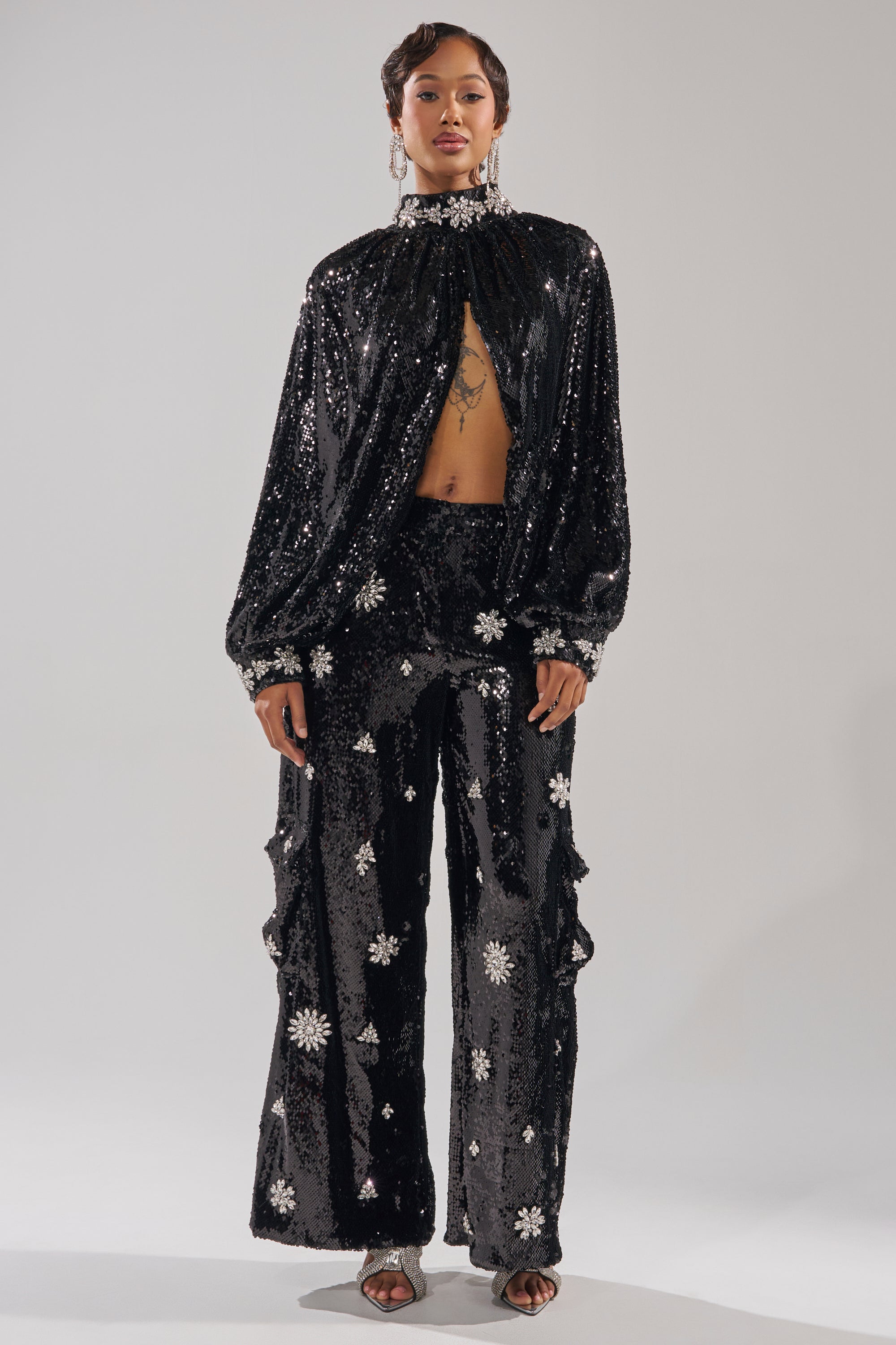 A woman wears the DAZZLING HOLIDAY SEQUIN WIDE LEG CARGO PANT paired with a high-neck, long-sleeve embellished top featuring silver floral details and a keyhole opening, styled with earrings and open-toe heels. 6