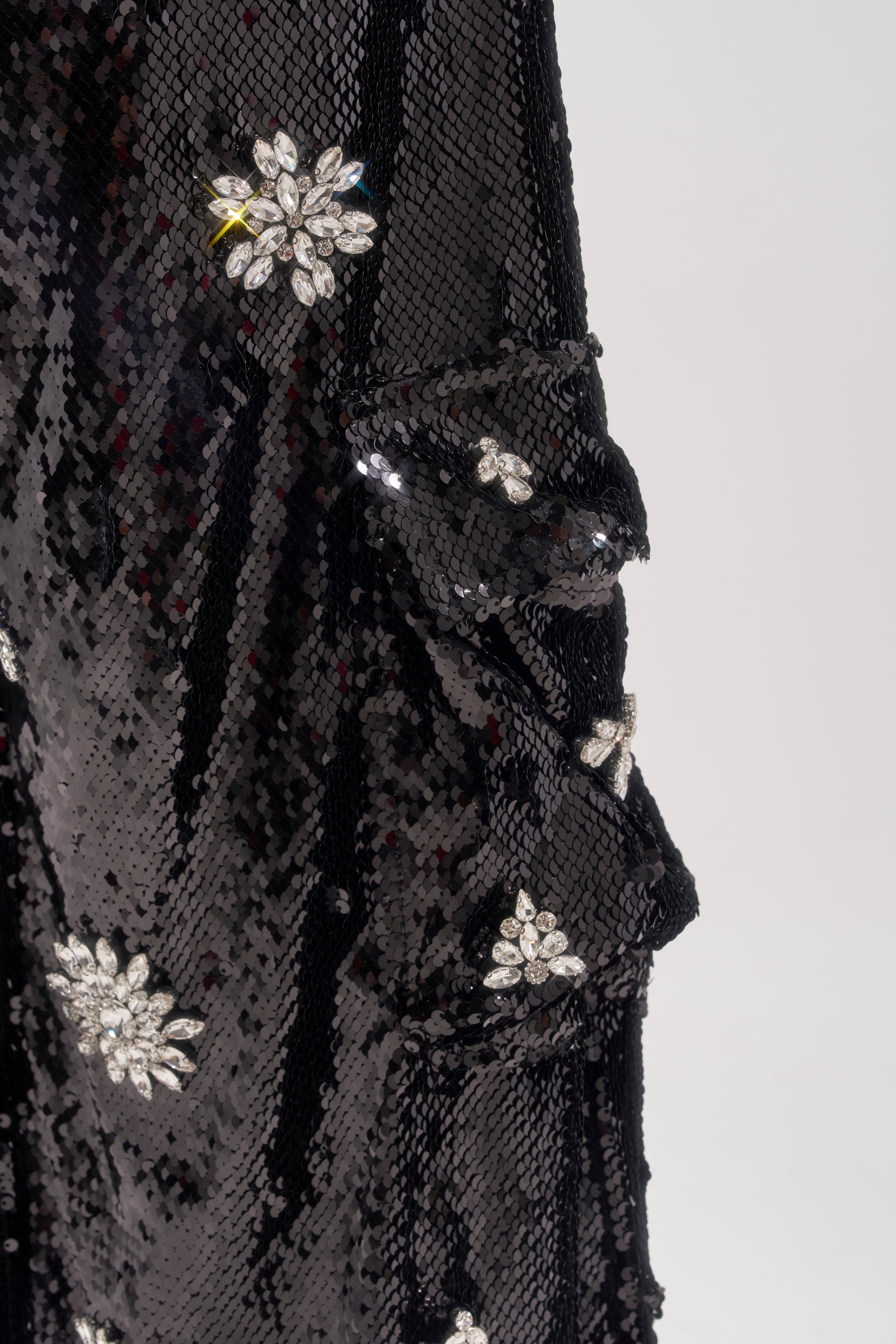 Close-up of black sequined fabric from the DAZZLING HOLIDAY SEQUIN WIDE LEG CARGO PANT, decorated with sparkling flower-shaped rhinestone accents and a touch of yellow embroidery on one flower, set against a plain light background. 7