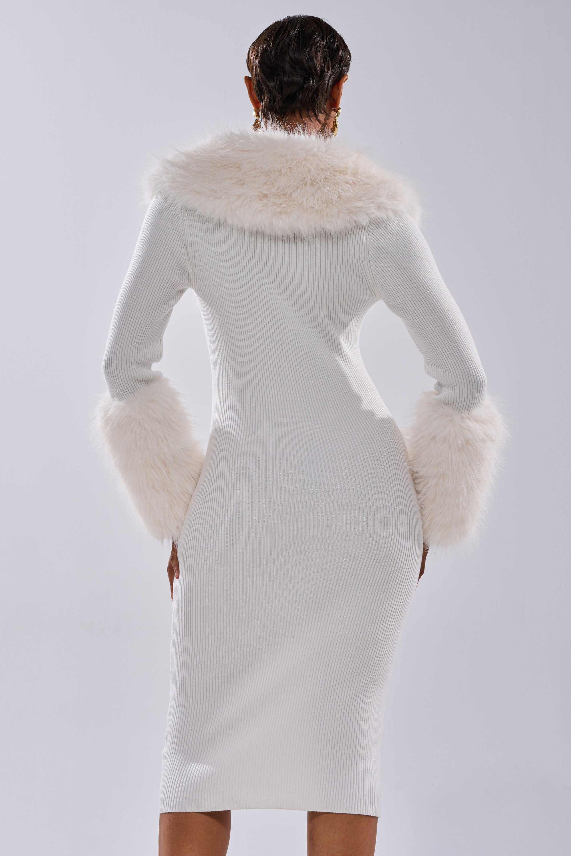 A person stands facing away, wearing the CELINE SWEATER MIDI DRESS WITH FUR TRIM—a fitted, ribbed knit dress featuring faux fur on the cuffs and collar. The background is plain and light-colored.