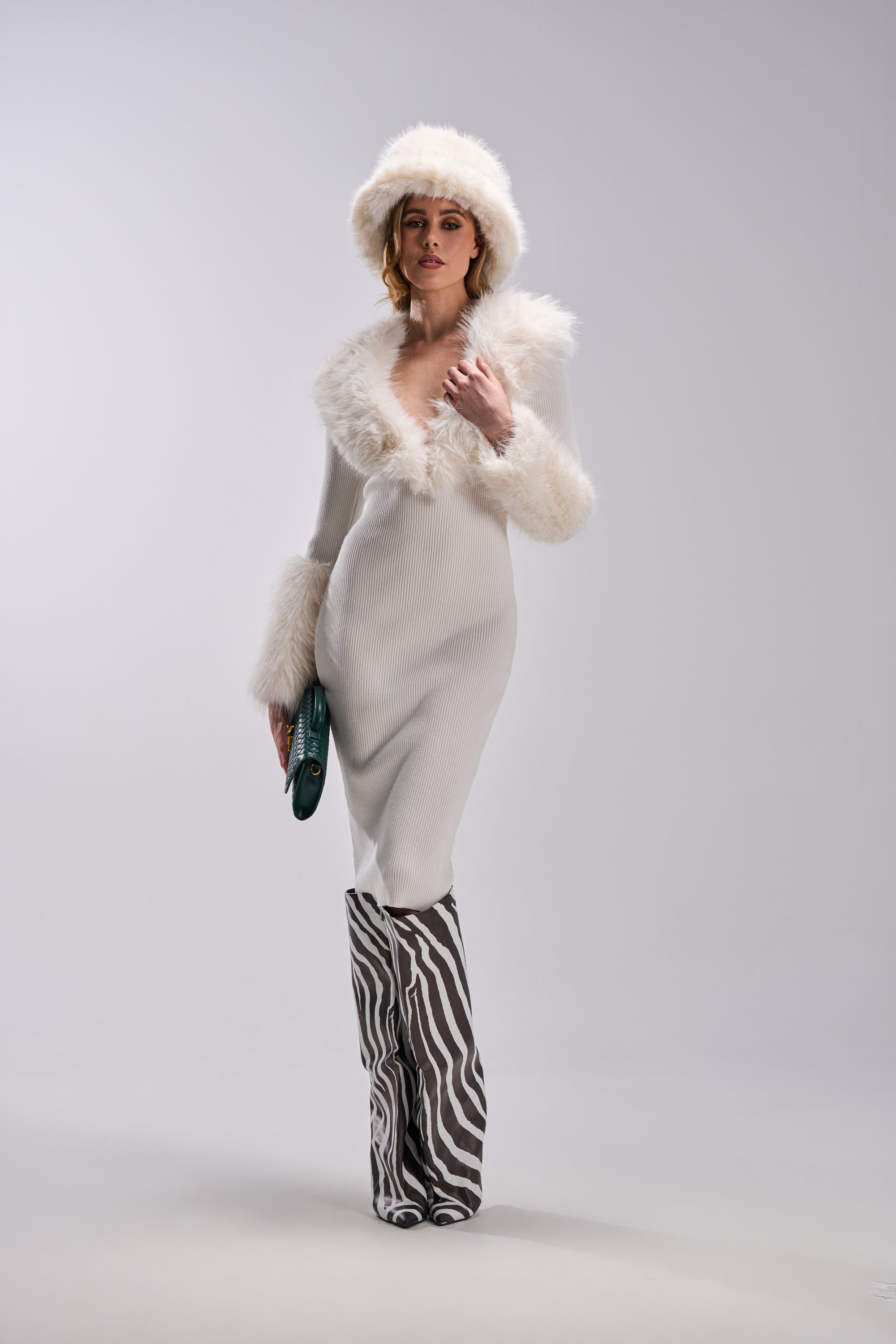 A woman wearing the CELINE SWEATER MIDI DRESS WITH FUR TRIM, a white fur hat, tall zebra-print boots, and holding a green clutch poses against a plain light background.