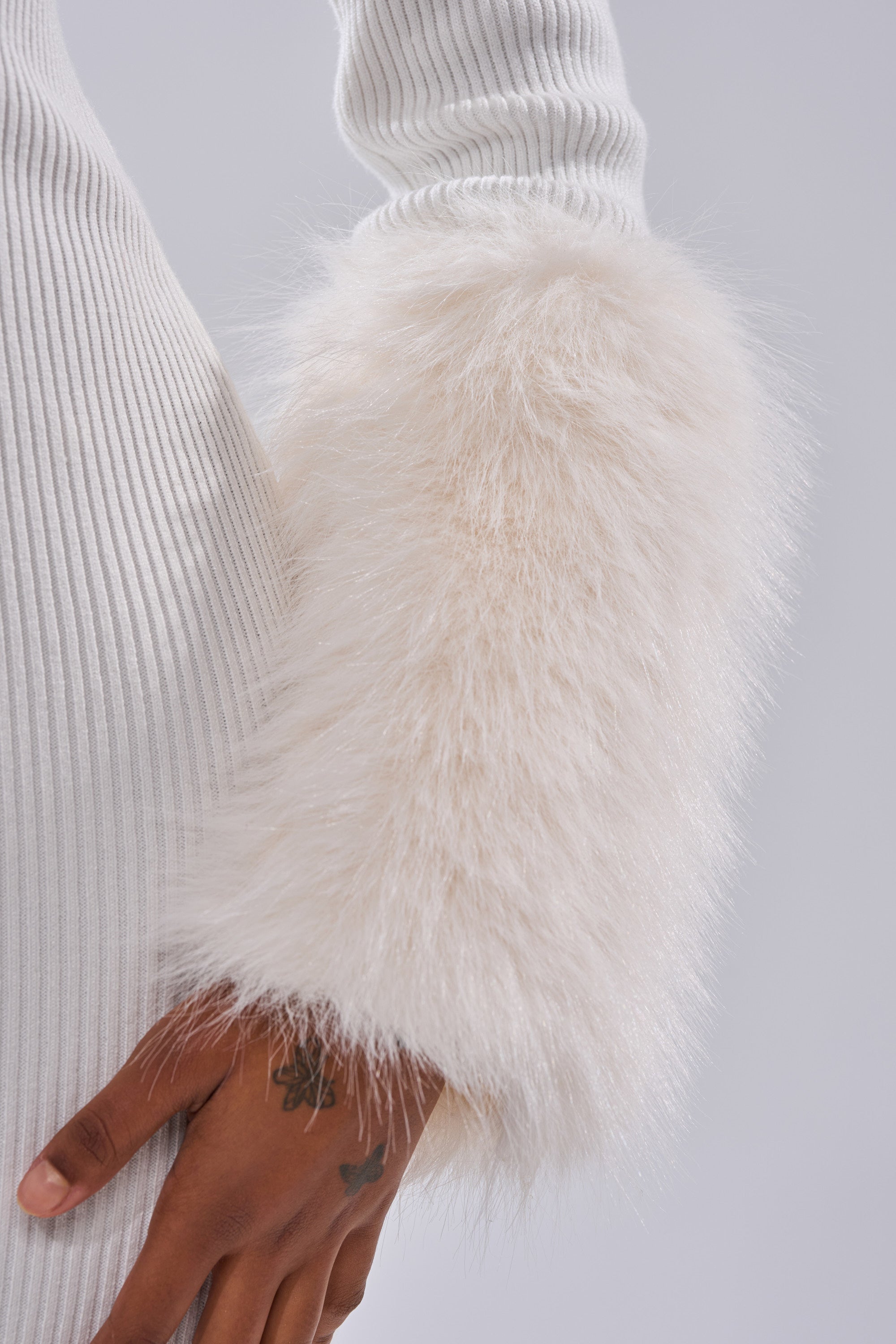 A close-up shows a person wearing the CELINE SWEATER MIDI DRESS WITH FUR TRIM, featuring ribbed knit fabric and large fluffy faux fur cuffs. Their tattooed wrist rests beside the dress against a light gray background.