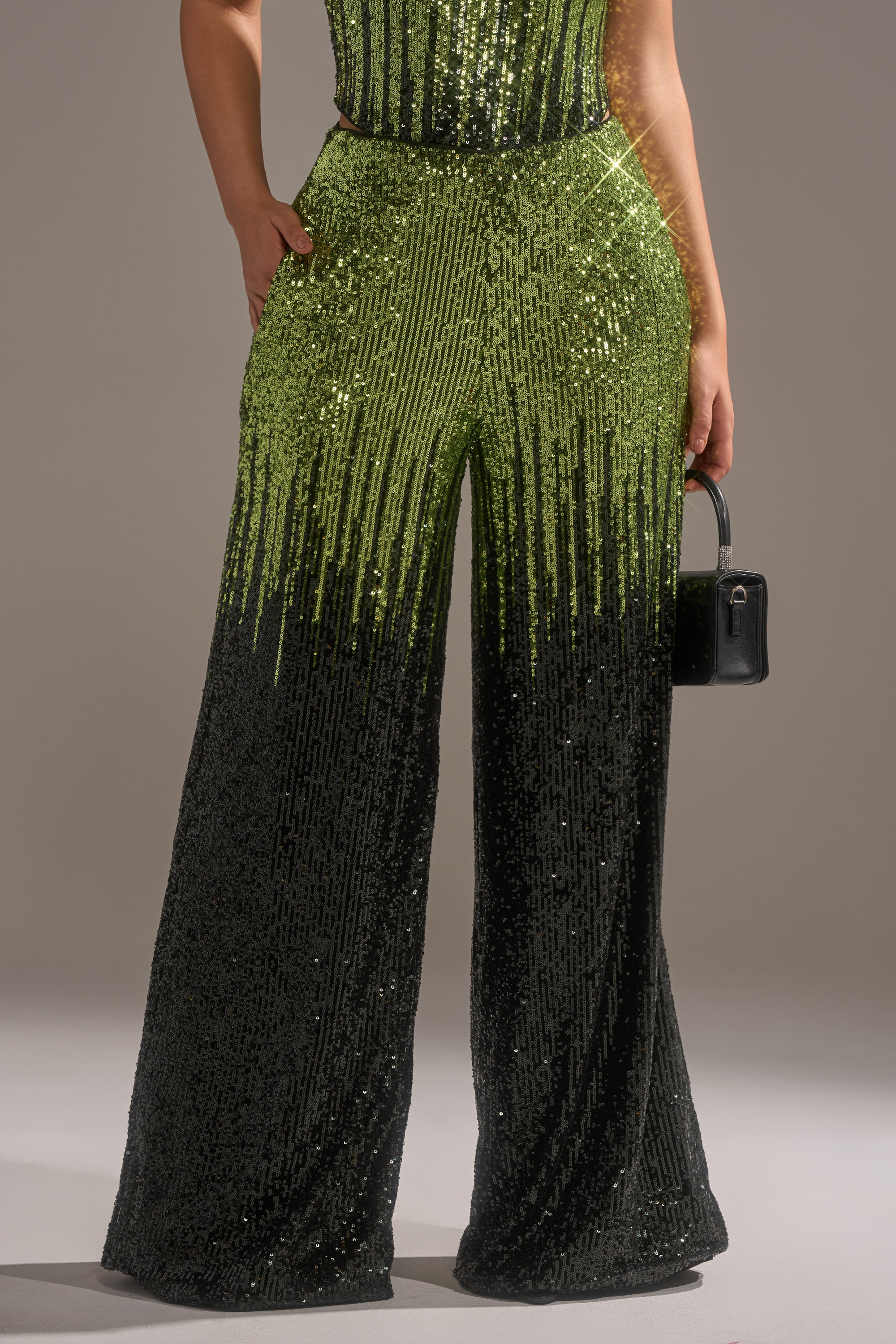 A person wearing the SWEET THING SEQUIN PALAZZO PANT, high-rise with a wide-leg silhouette and a bright green to black sequin gradient, holding a small black handbag.