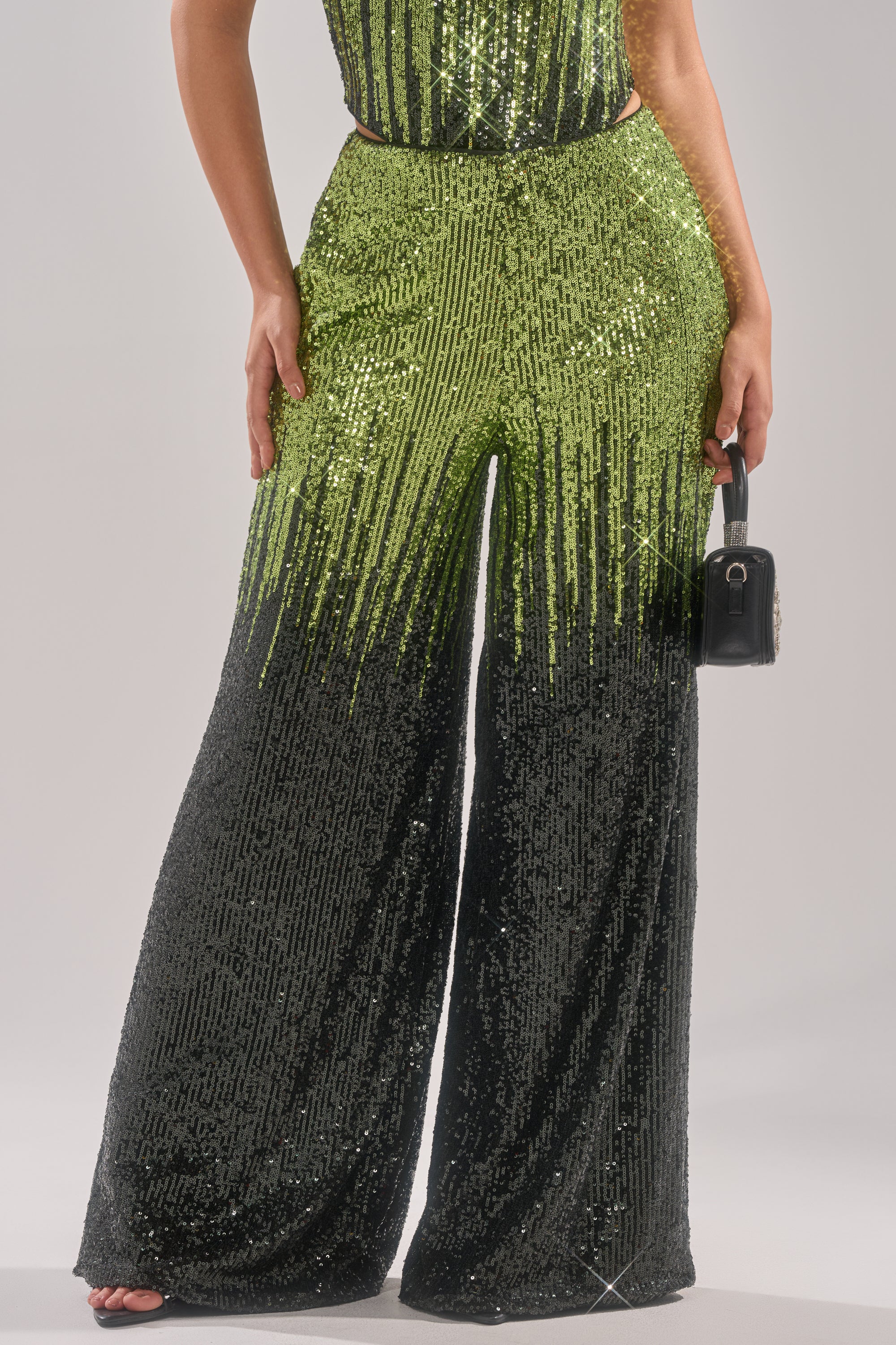 Wearing the SWEET THING SEQUIN PALAZZO PANT—high-rise, wide-leg with green-to-black sequins—a person stands against a plain background holding a small black handbag.