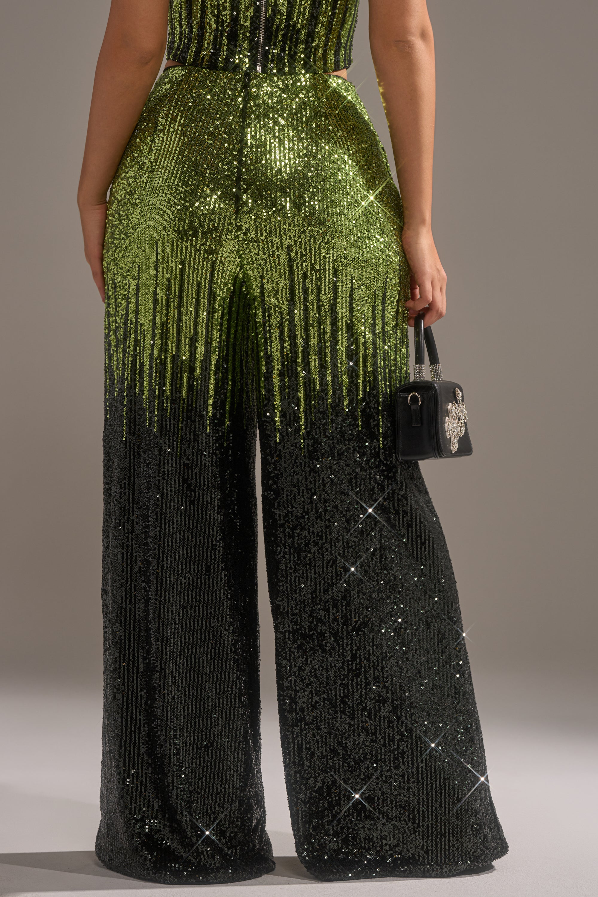 Someone stands facing away, wearing the SWEET THING SEQUIN PALAZZO PANT in green and black sequins, and holds a small black handbag adorned with jeweled embellishments.
