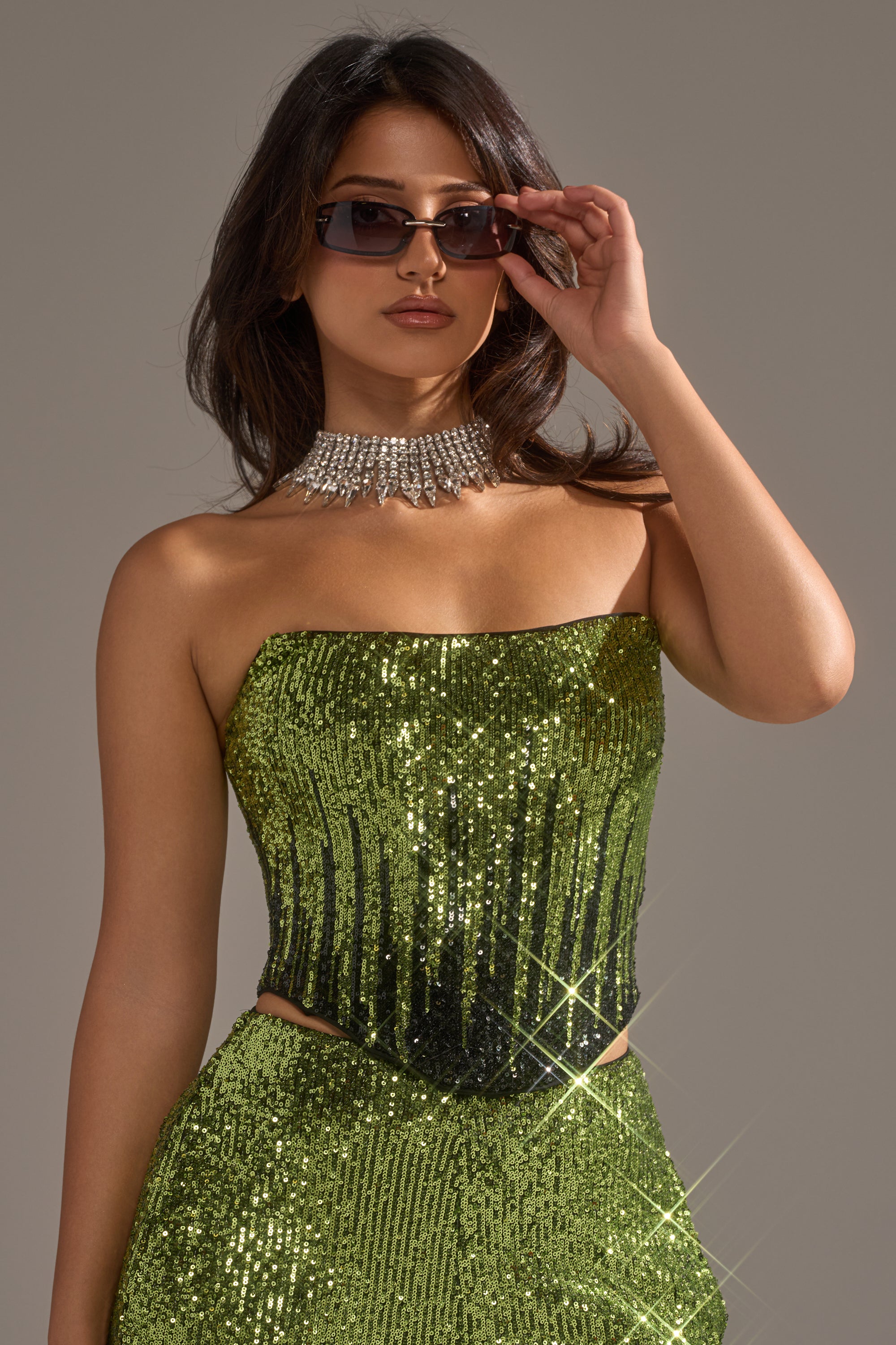 A woman confidently poses against a neutral background in the SWEET THING SEQUIN CORSET paired with a matching skirt, statement rhinestone necklace, and dark sunglasses.