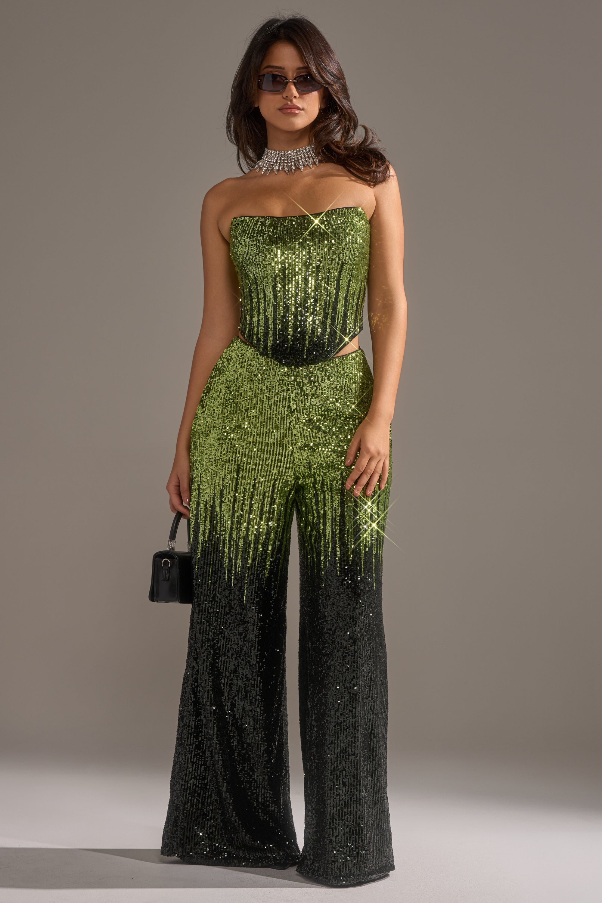 A woman wearing sunglasses, the SWEET THING SEQUIN CORSET—a strapless green and black ombre sequin jumpsuit—with a rhinestone choker and small black handbag stands against a plain background.