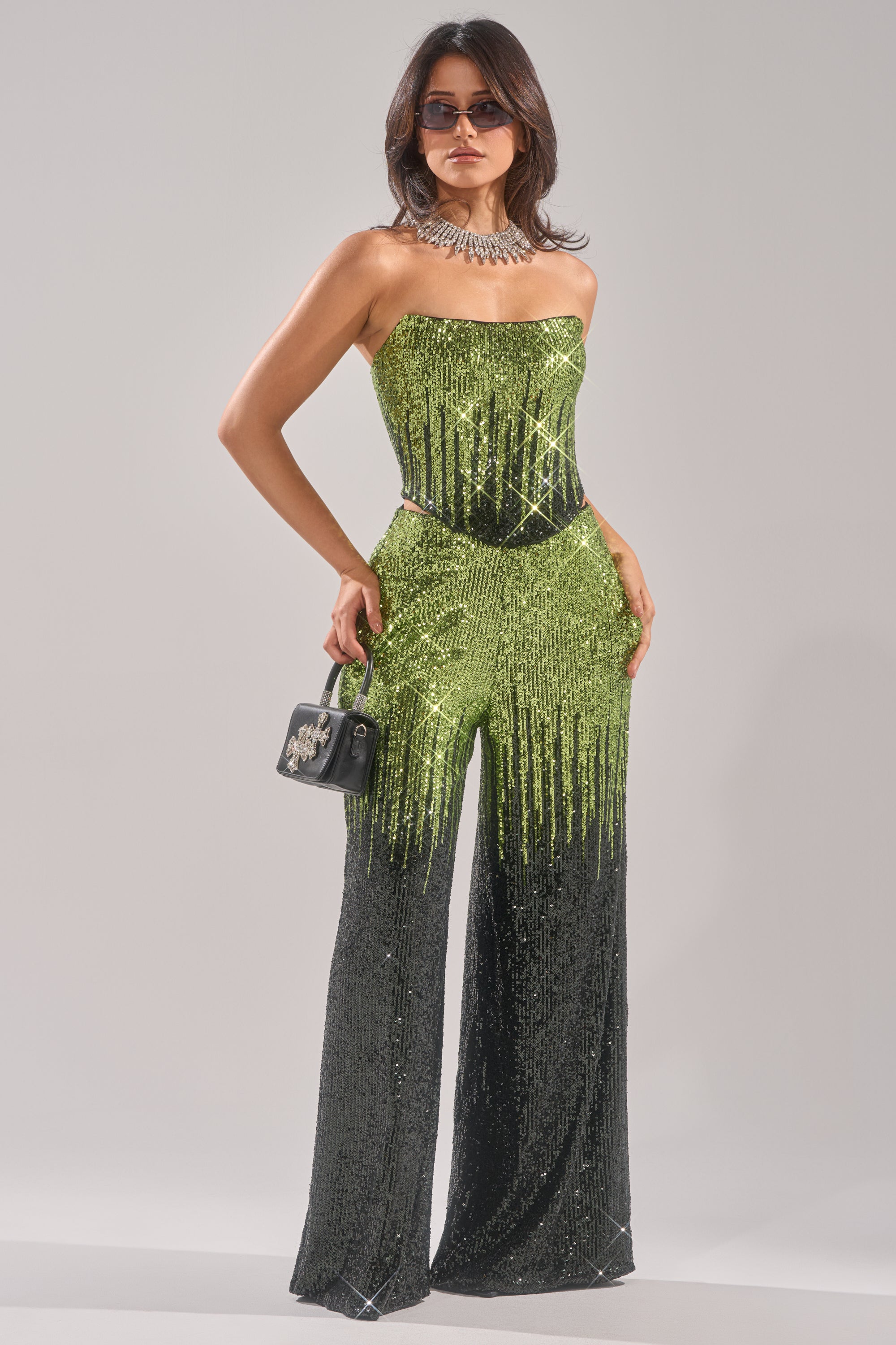 A woman wears the SWEET THING SEQUIN CORSET, a strapless ombre green jumpsuit fading to black, styled with sunglasses, a chunky necklace, and a small black handbag.