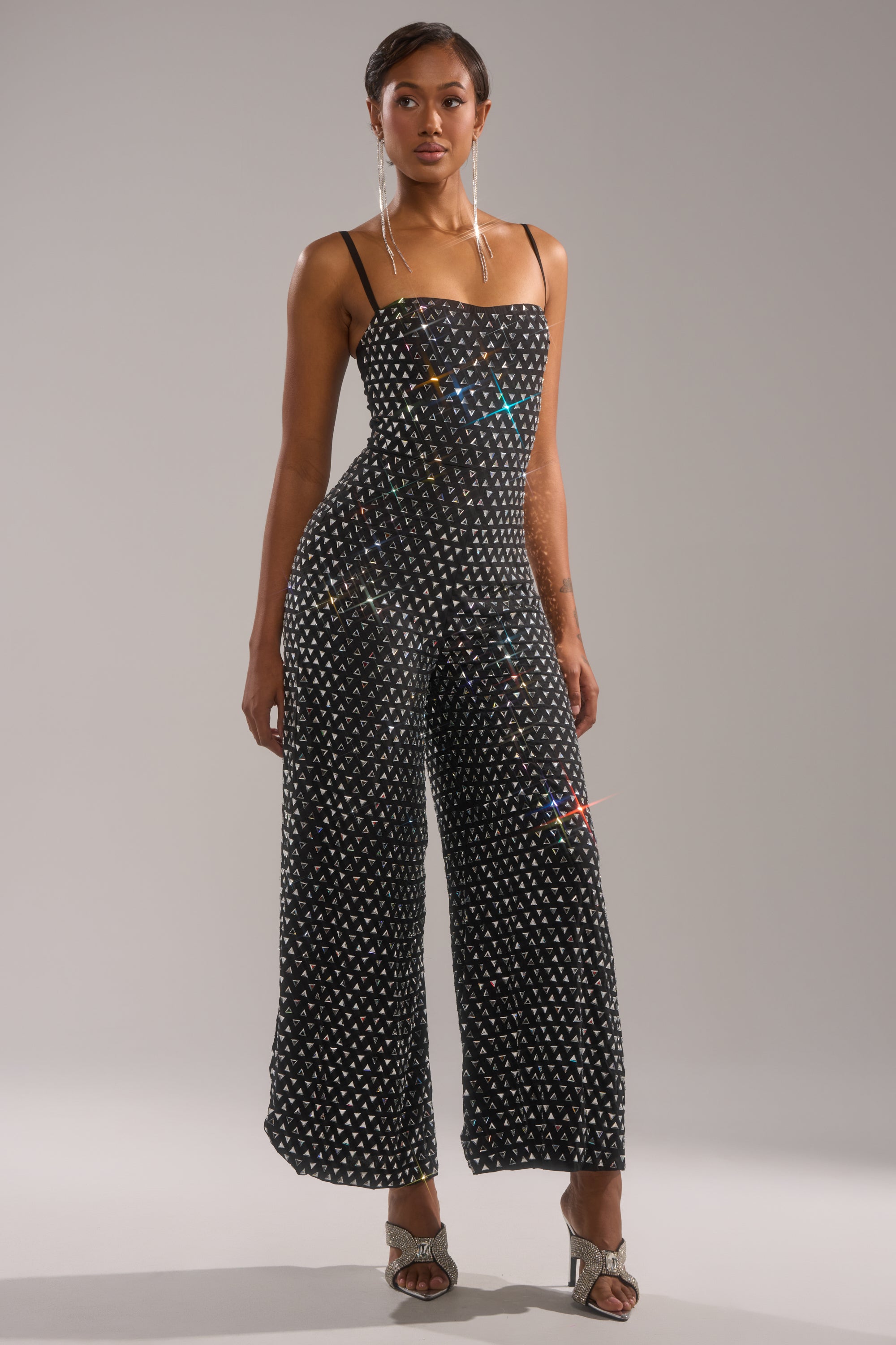 A woman exudes confidence in the CLARA RHINESTONE WIDE LEG JUMPSUIT, a black party piece with a bold white dot design. She completes her look with statement earrings and strappy metallic heels against a plain gray background.