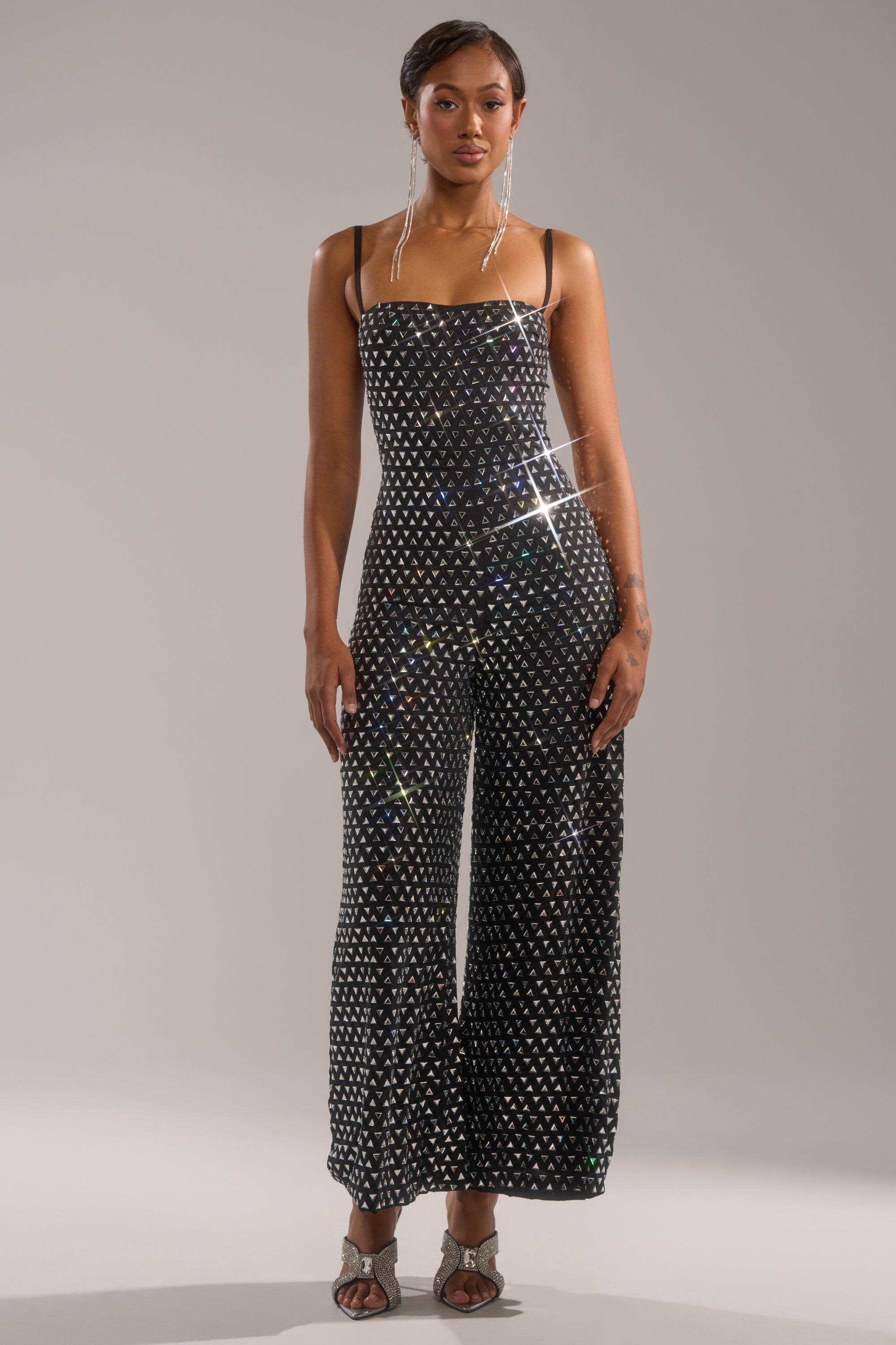A woman faces forward in the CLARA RHINESTONE WIDE LEG JUMPSUIT, featuring black fabric and sparkling rhinestones. She pairs it with earrings and sandals, her short hair styled sleekly against a plain gray background.