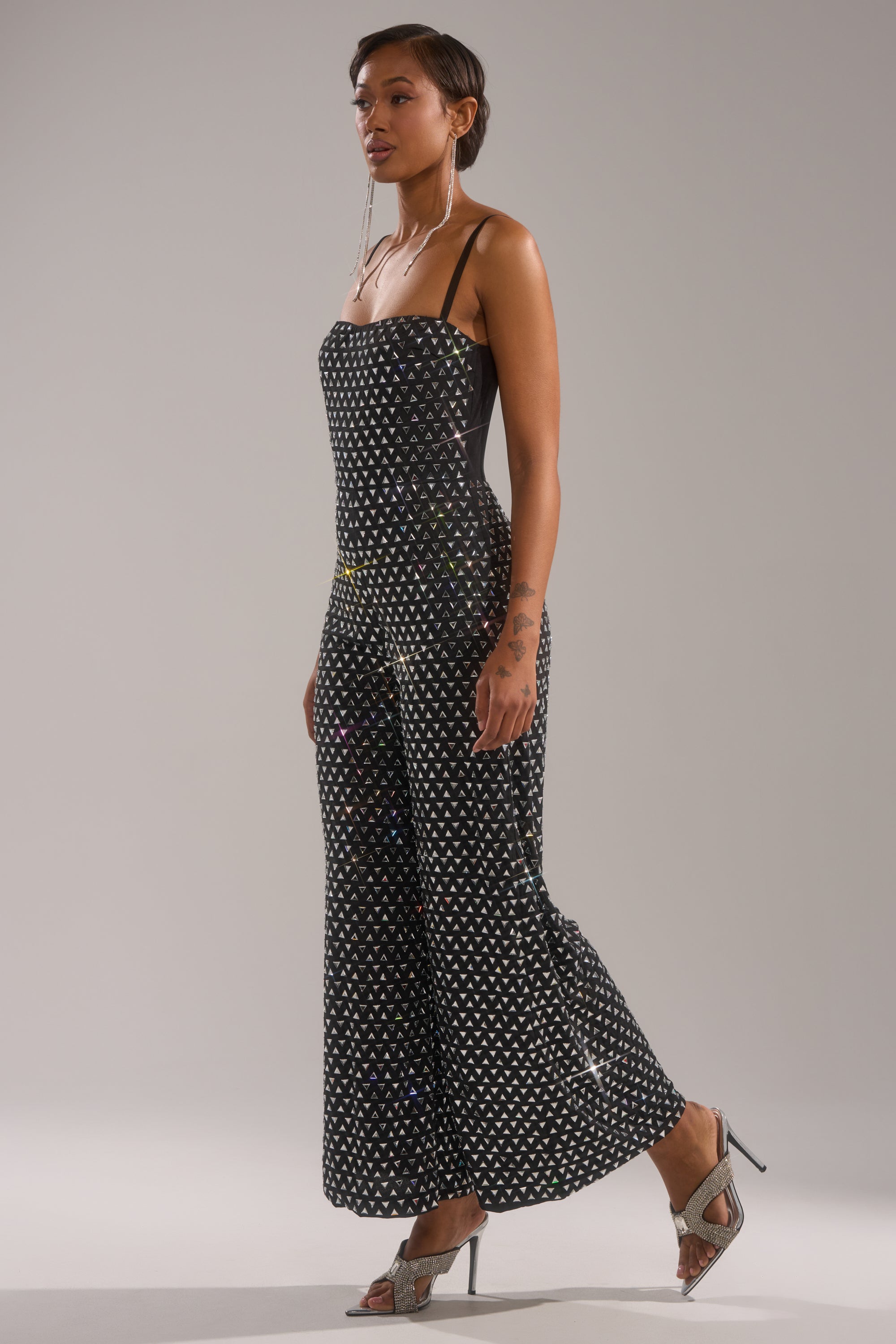 A woman in profile models the CLARA RHINESTONE WIDE LEG JUMPSUIT, a black strapless wide-leg style accented with white polka dots. She wears dangling earrings, high-heeled sandals, and displays tattoos on her arm against a plain background.
