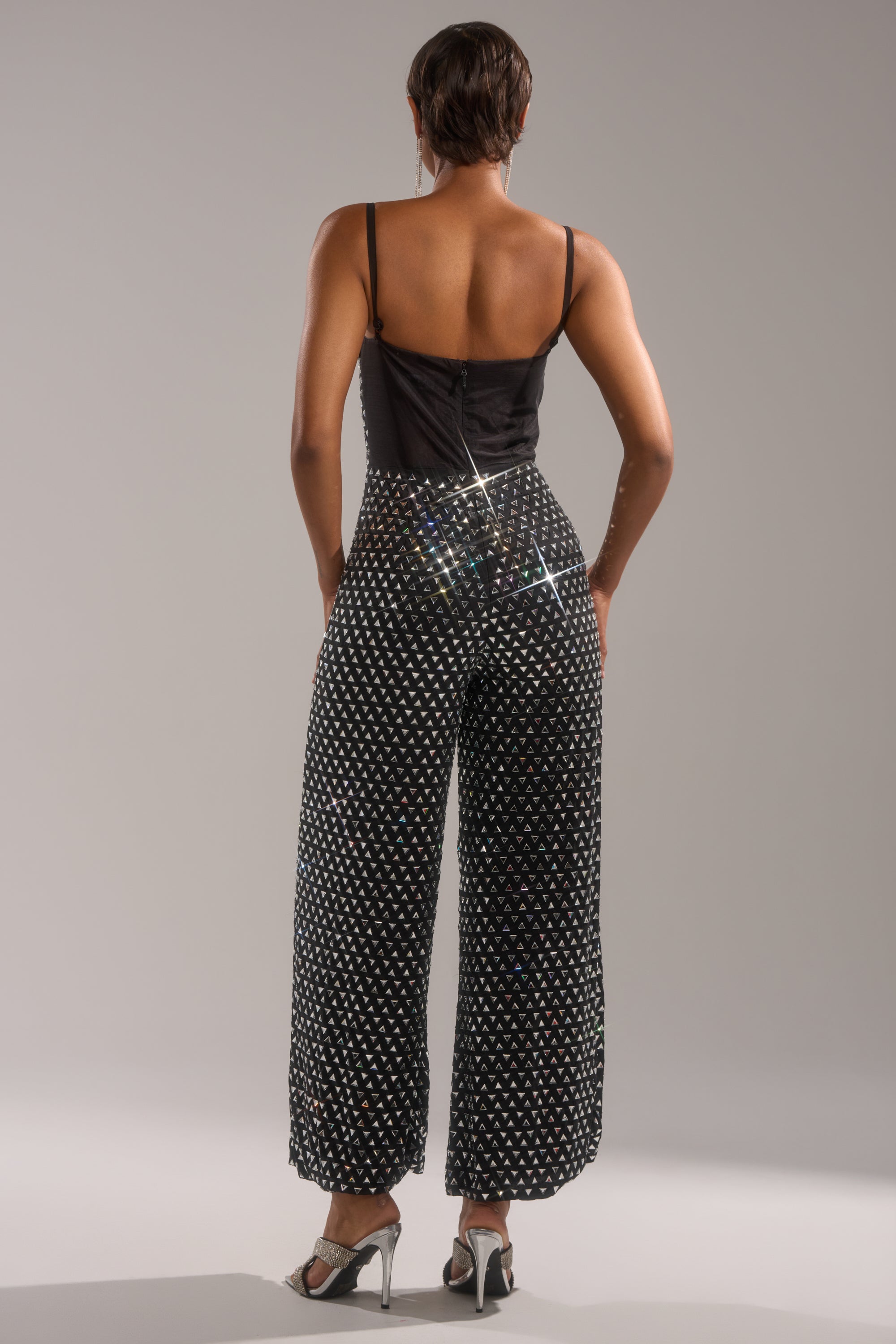 A person stands with their back to the camera, wearing the CLARA RHINESTONE WIDE LEG JUMPSUIT—sleeveless, black, with a white dotted pattern—and high heels, set against a plain light background.
