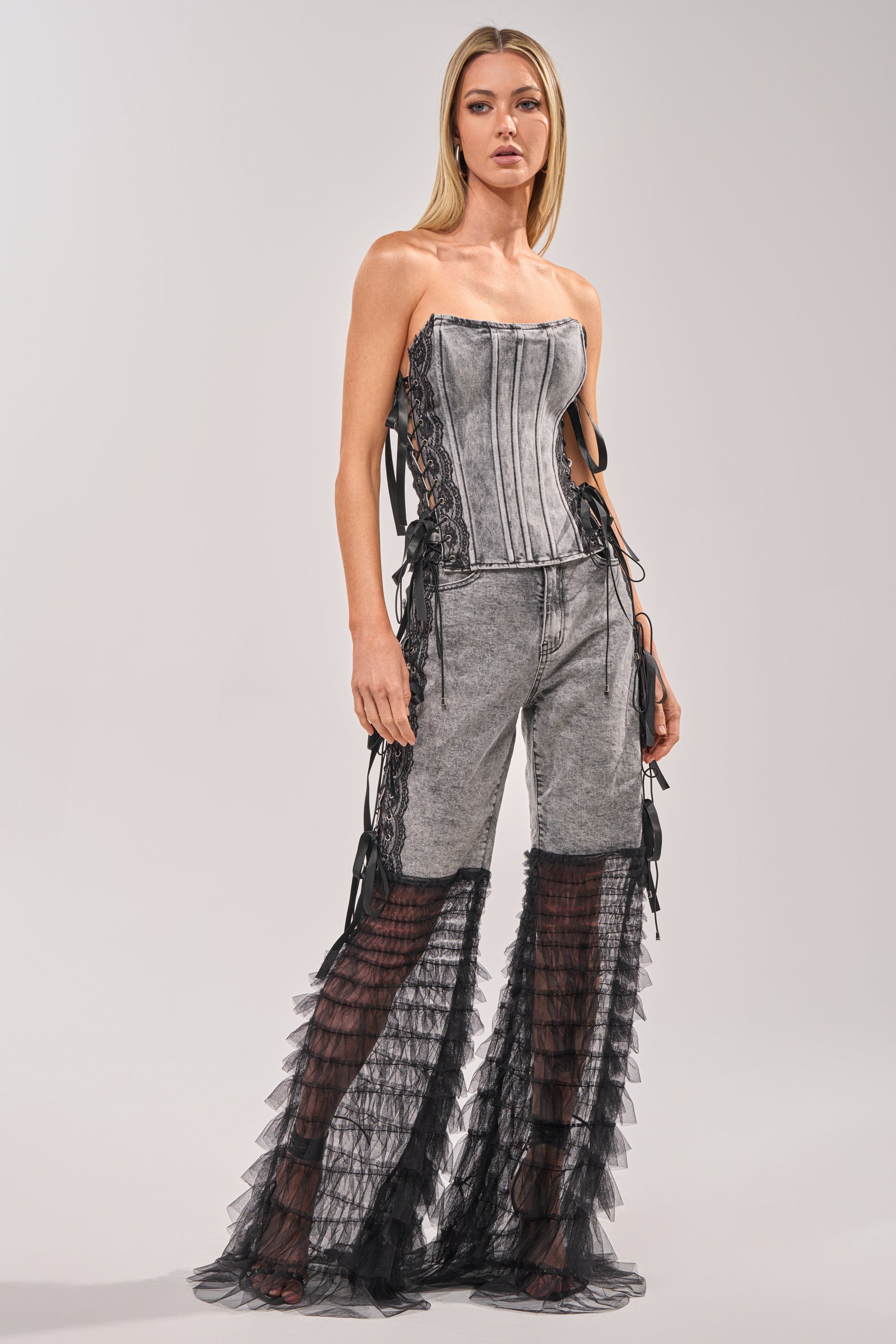 A woman stands against a plain light background wearing the TICKED AND TIED LACE DETAIL FLARE DENIM, styled with a gray, strapless corset top and lace-up sides, paired with flare pants featuring sheer black ruffle details from knee to hem. 0