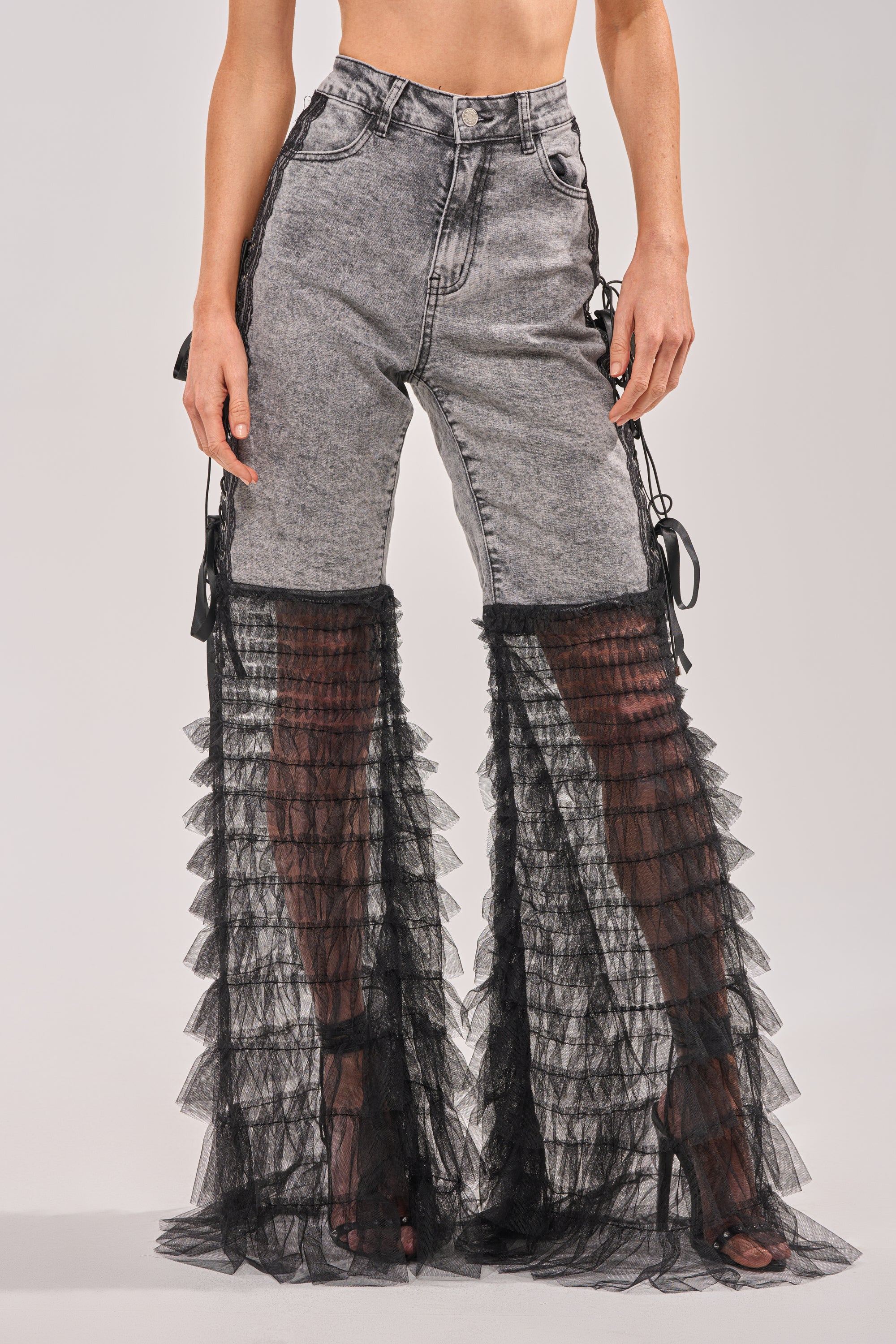 A person wears the TICKED AND TIED LACE DETAIL FLARE DENIM, high-waisted gray acid-wash jeans with sheer black ruffled tulle from knee to ankle, paired with black heels for a chic, fitted look. 2