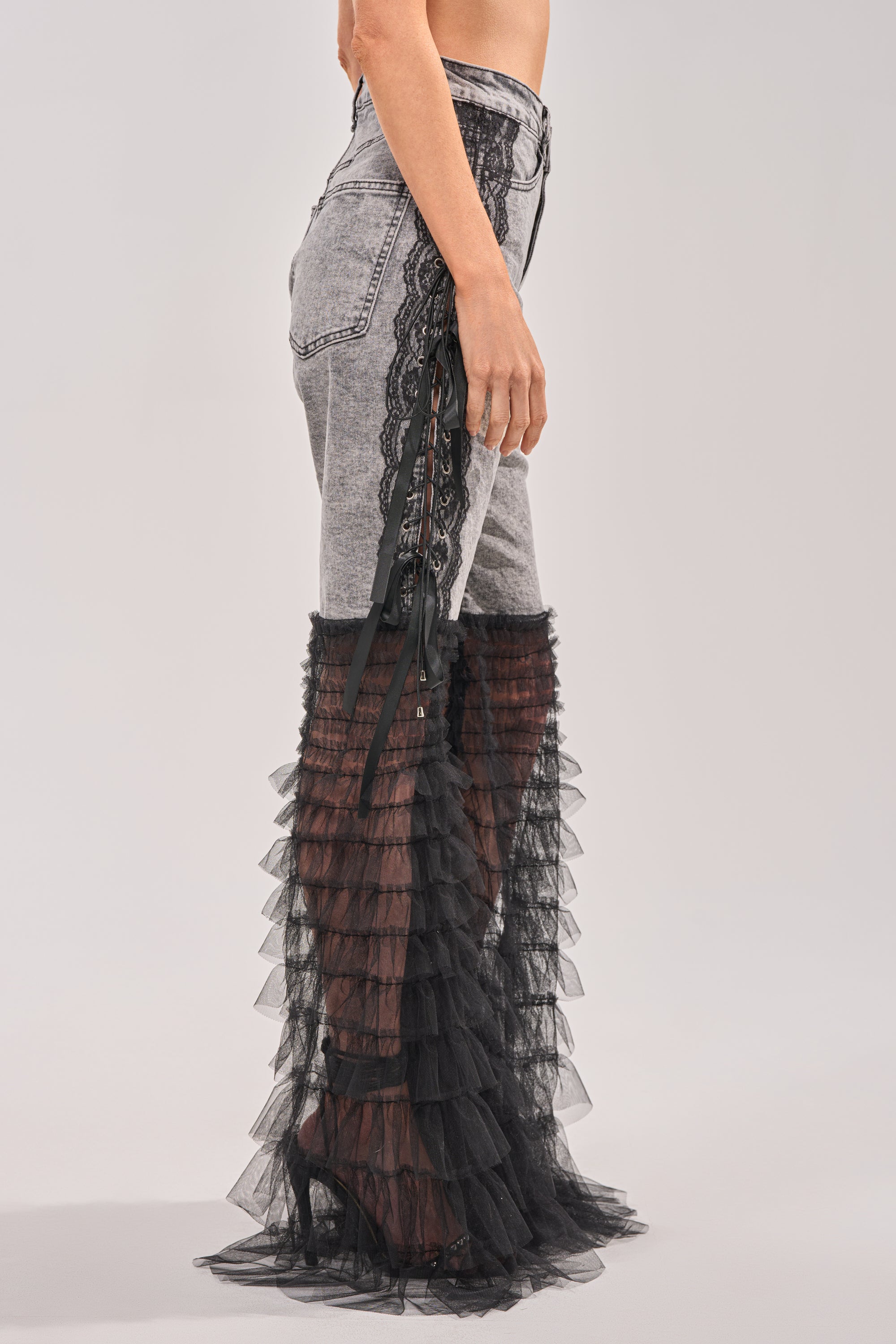Someone wearing the TICKED AND TIED LACE DETAIL FLARE DENIM, featuring high-rise light gray jeans with black lace details and sheer tiered tulle overlays from thigh to ankle, styled with high-heeled shoes. 3