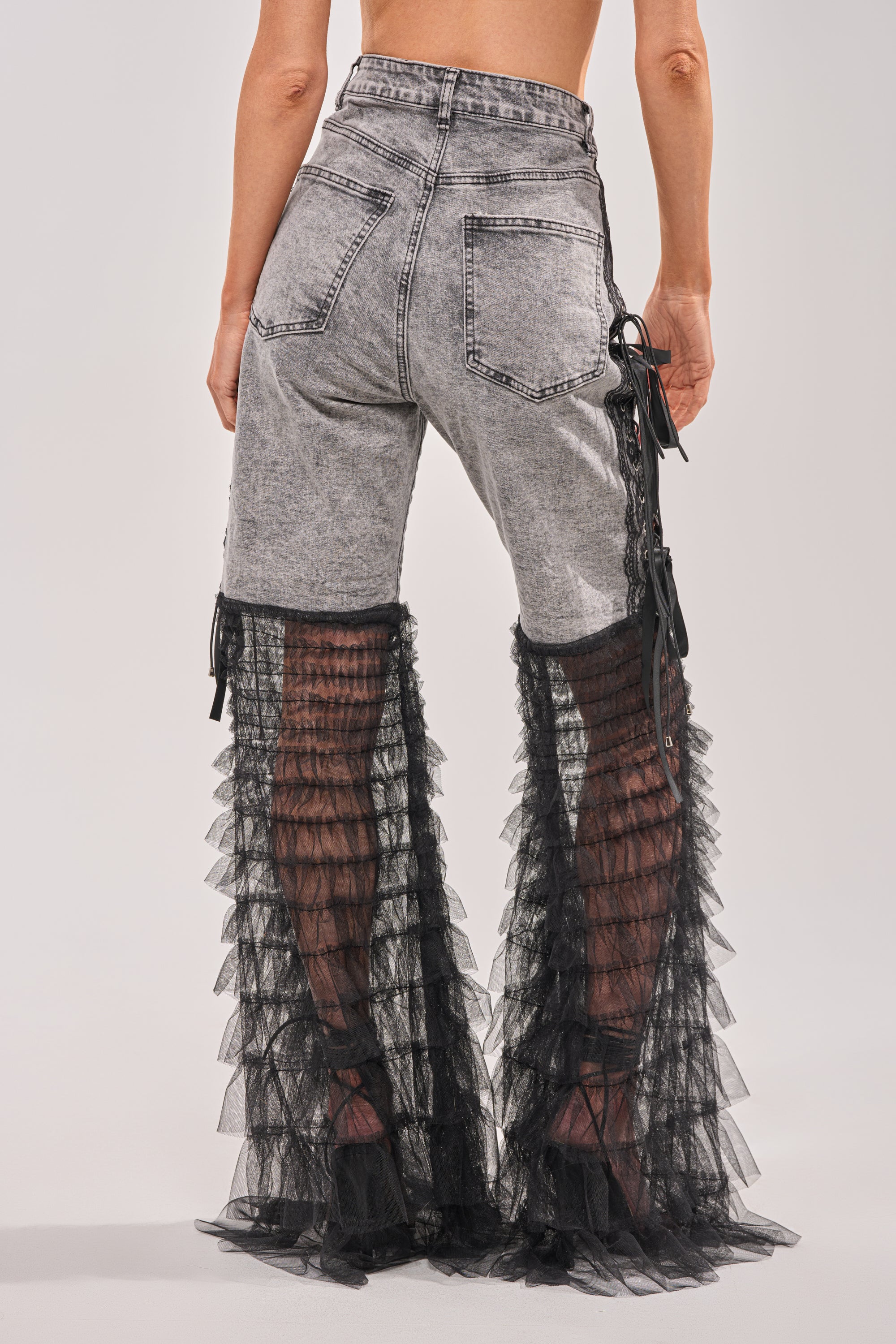 A person stands barefoot, facing away on a plain backdrop, wearing the TICKED AND TIED LACE DETAIL FLARE DENIM—high-rise gray jeans featuring sheer black tulle ruffles and delicate lace accents from knee to hem. 4