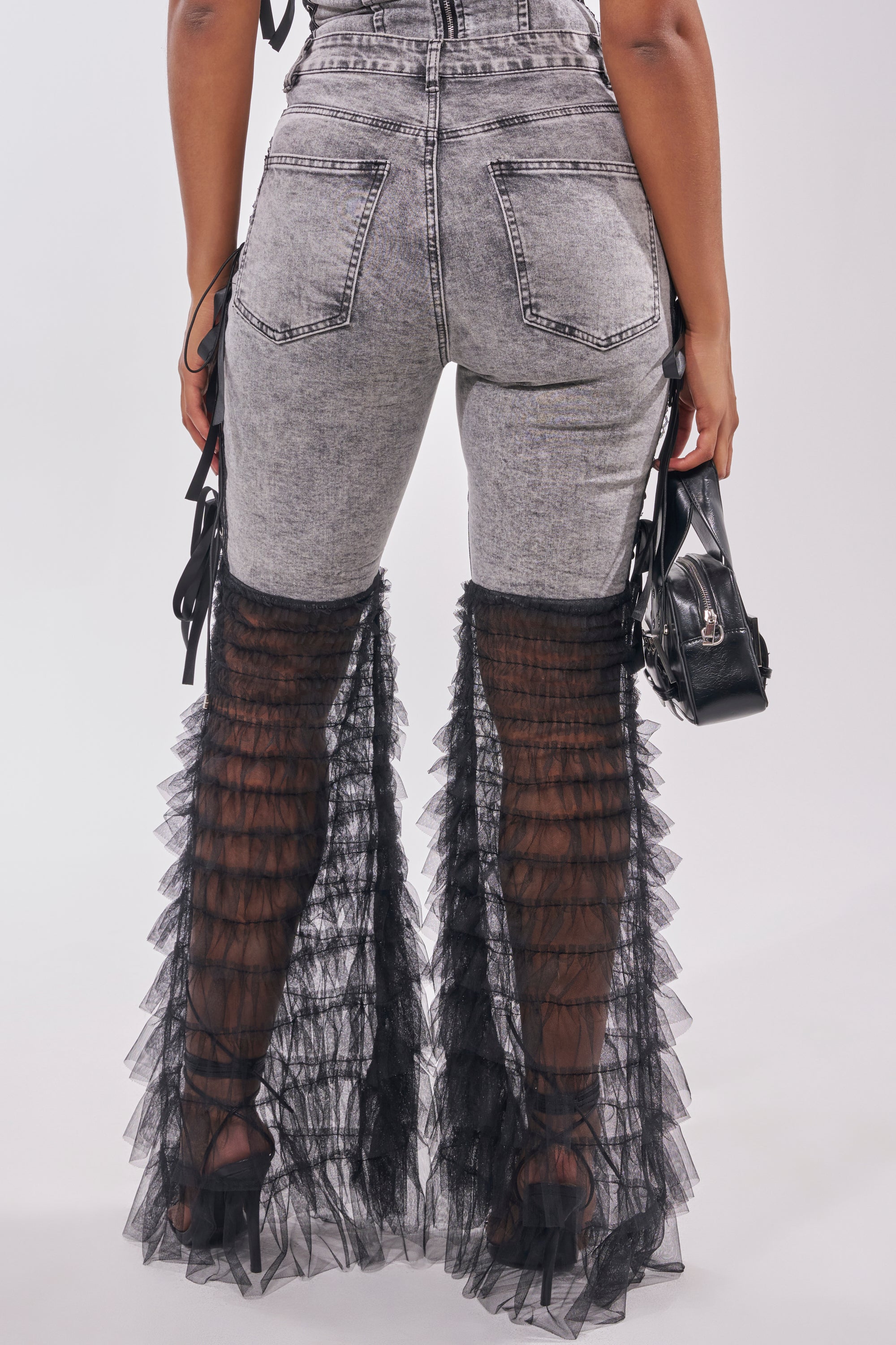 A person is shown from behind wearing TICKED AND TIED LACE DETAIL FLARE DENIM with black sheer ruffles over the lower legs, paired with black heels and a small black handbag, against a plain background. 4