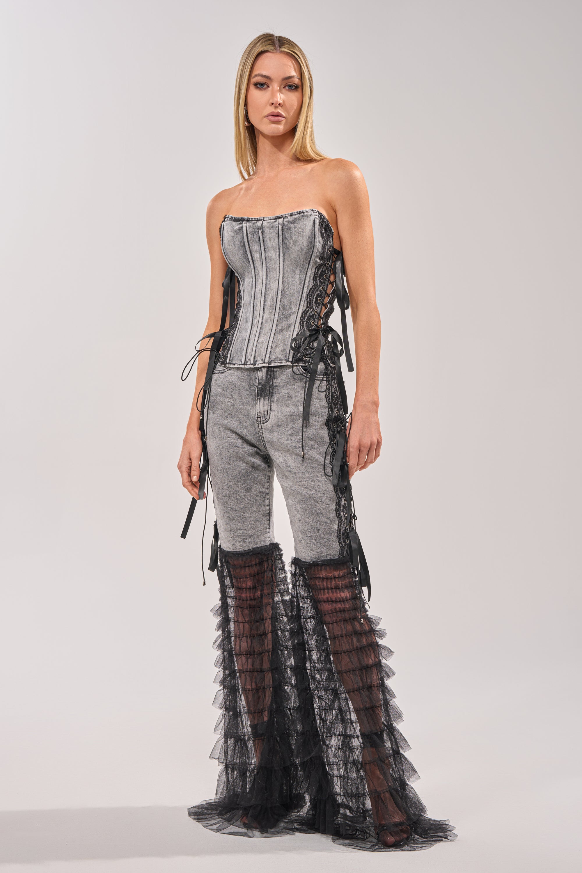 A woman poses against a plain light background, wearing the TICKED AND TIED LACE DETAIL FLARE DENIM—a gray strapless corset top paired with high-rise jeans featuring sheer black ruffle panels from the knees down. 5