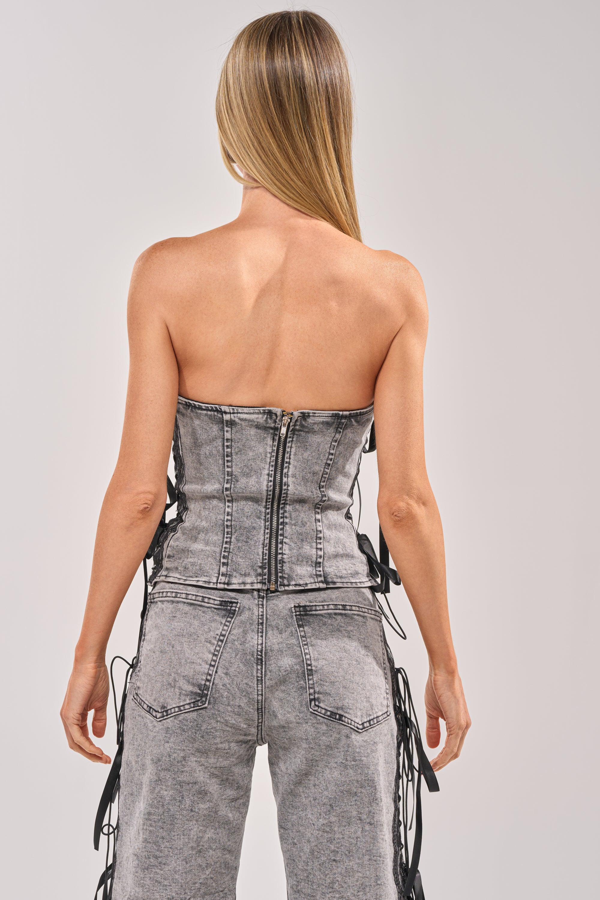 A woman with straight blonde hair wears the TICKED AND TIED LACE DETAIL DENIM CORSET and matching jeans, both in faded gray with black side ties and subtle lace details. She faces away, highlighting the back zipper and seams. 3