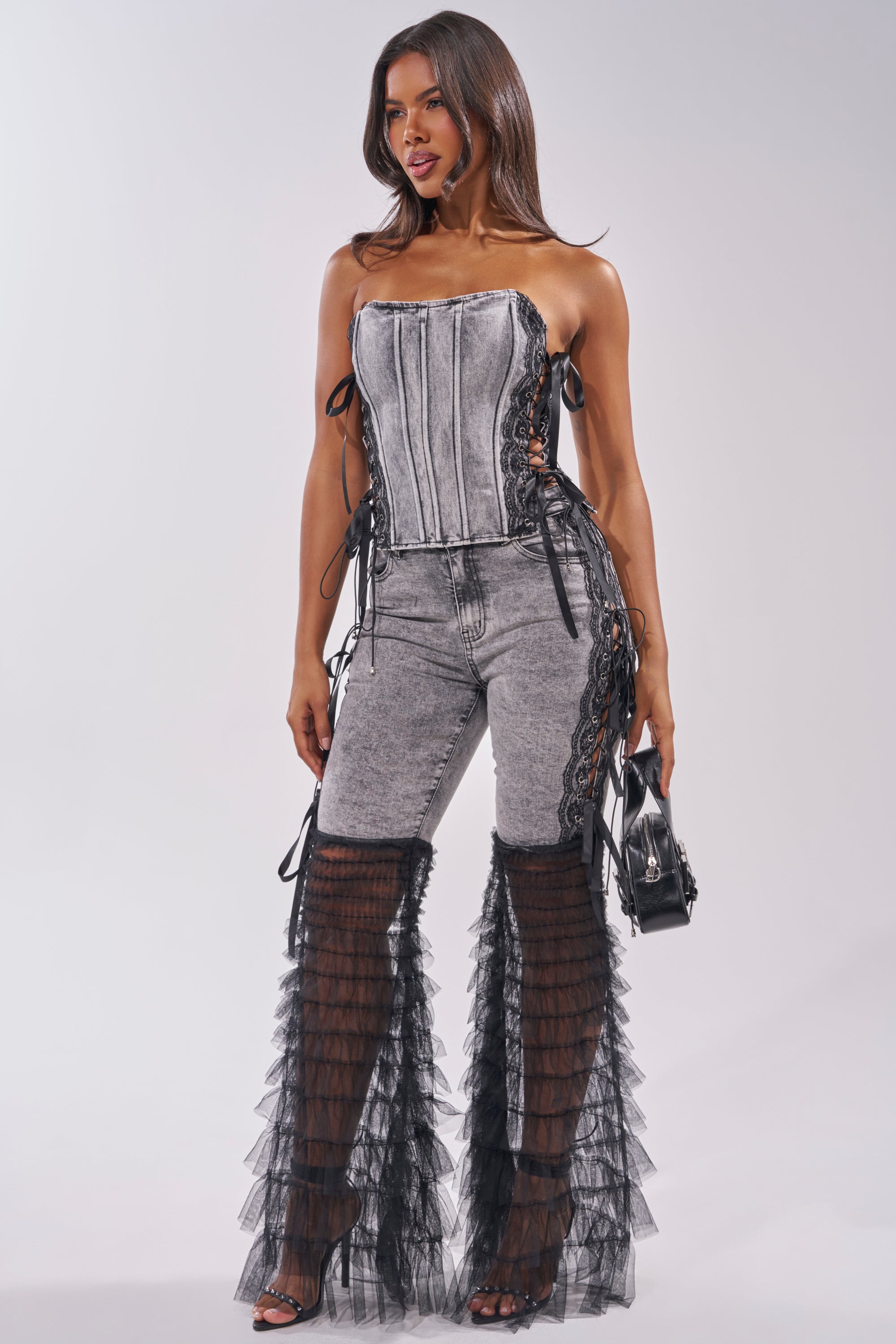 A woman poses against a light background in the TICKED AND TIED LACE DETAIL DENIM CORSET with distressed grey jeans featuring black lace-up details and sheer ruffled overlays, accessorized with a small black handbag and heeled sandals. 5