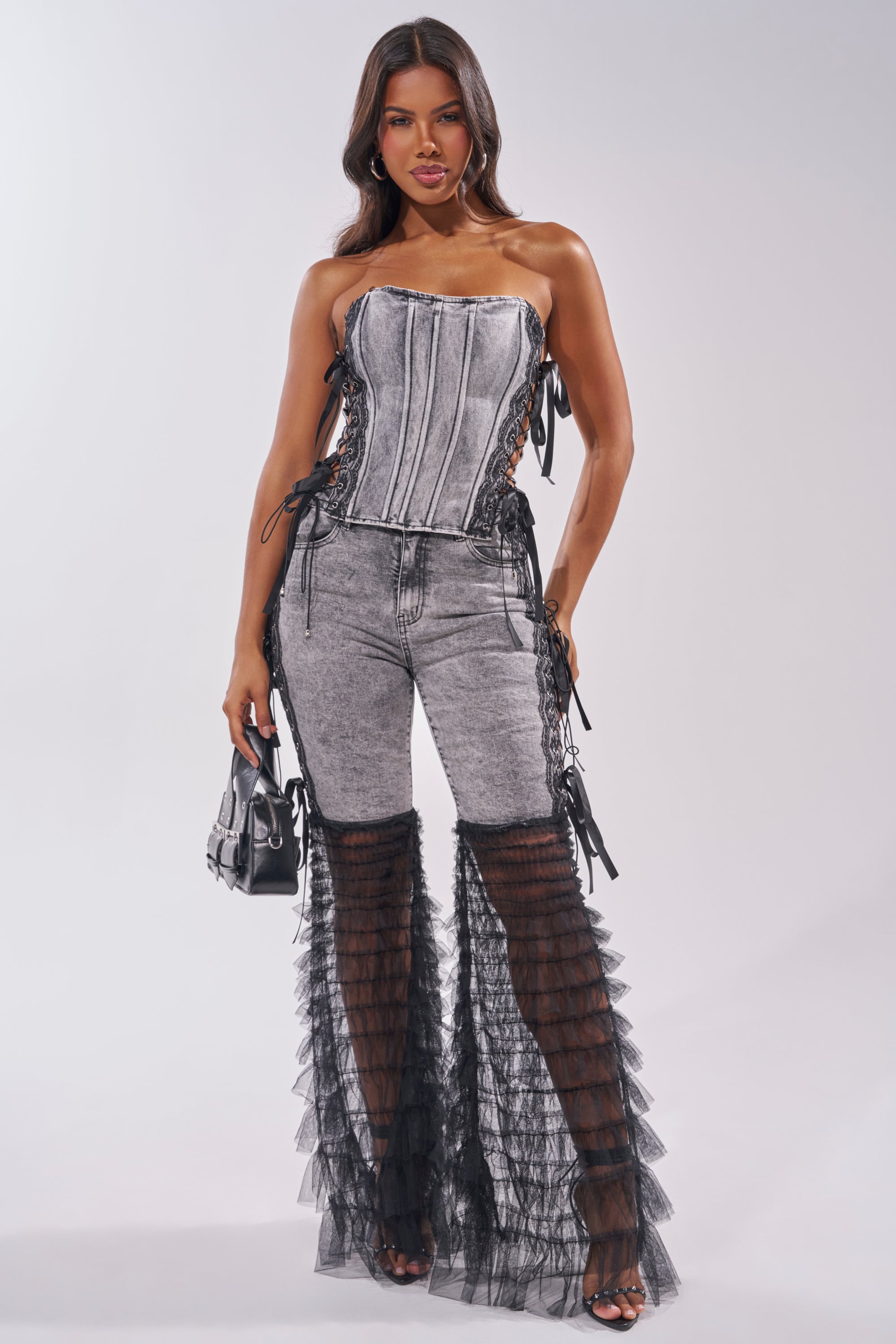 A woman stands confidently in the TICKED AND TIED LACE DETAIL DENIM CORSET, styled with fitted grey jeans featuring bold black sheer ruffled flares, a small black handbag, and black heels. 6