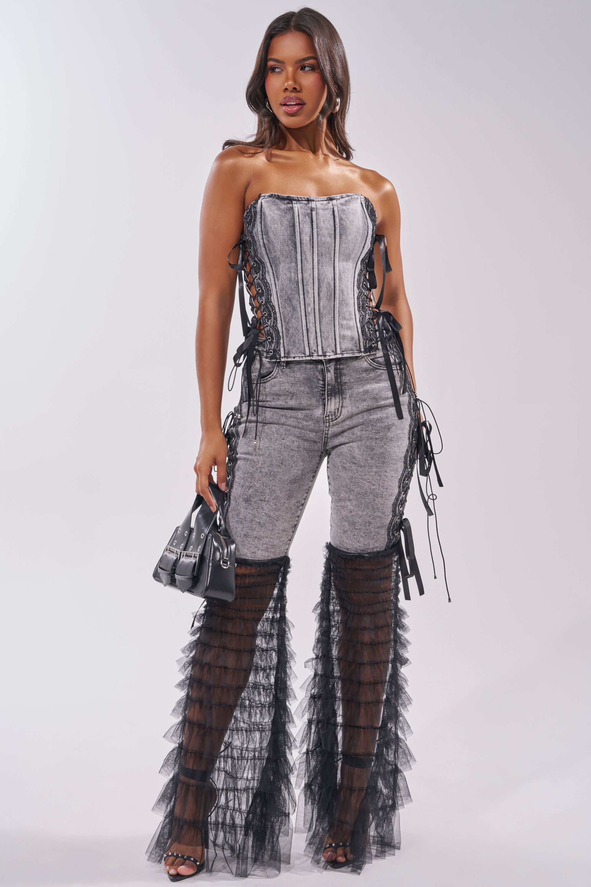 A woman stands confidently in the TICKED AND TIED LACE DETAIL DENIM CORSET, paired with high-waisted acid-wash jeans featuring lace satin side details, black sheer ruffled chaps, high heels, and a small black handbag against a light gray background. 7
