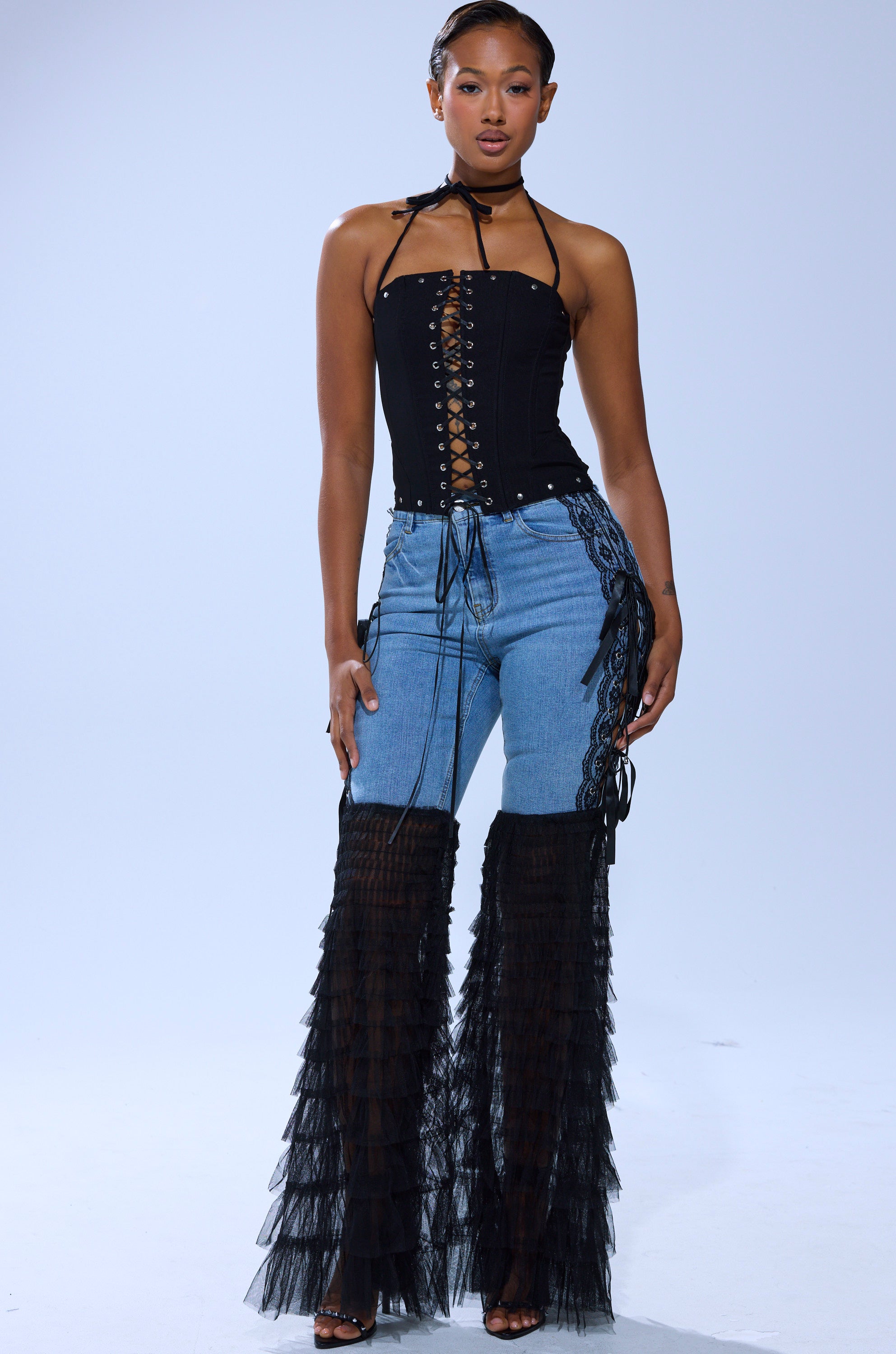 A person stands confidently in black high-heeled sandals and the CAN'T DECIDE TULLE AND LACE DENIM JEANS featuring a lace-up front and ruffled flare legs, paired with a black lace-up corset top against a plain background.