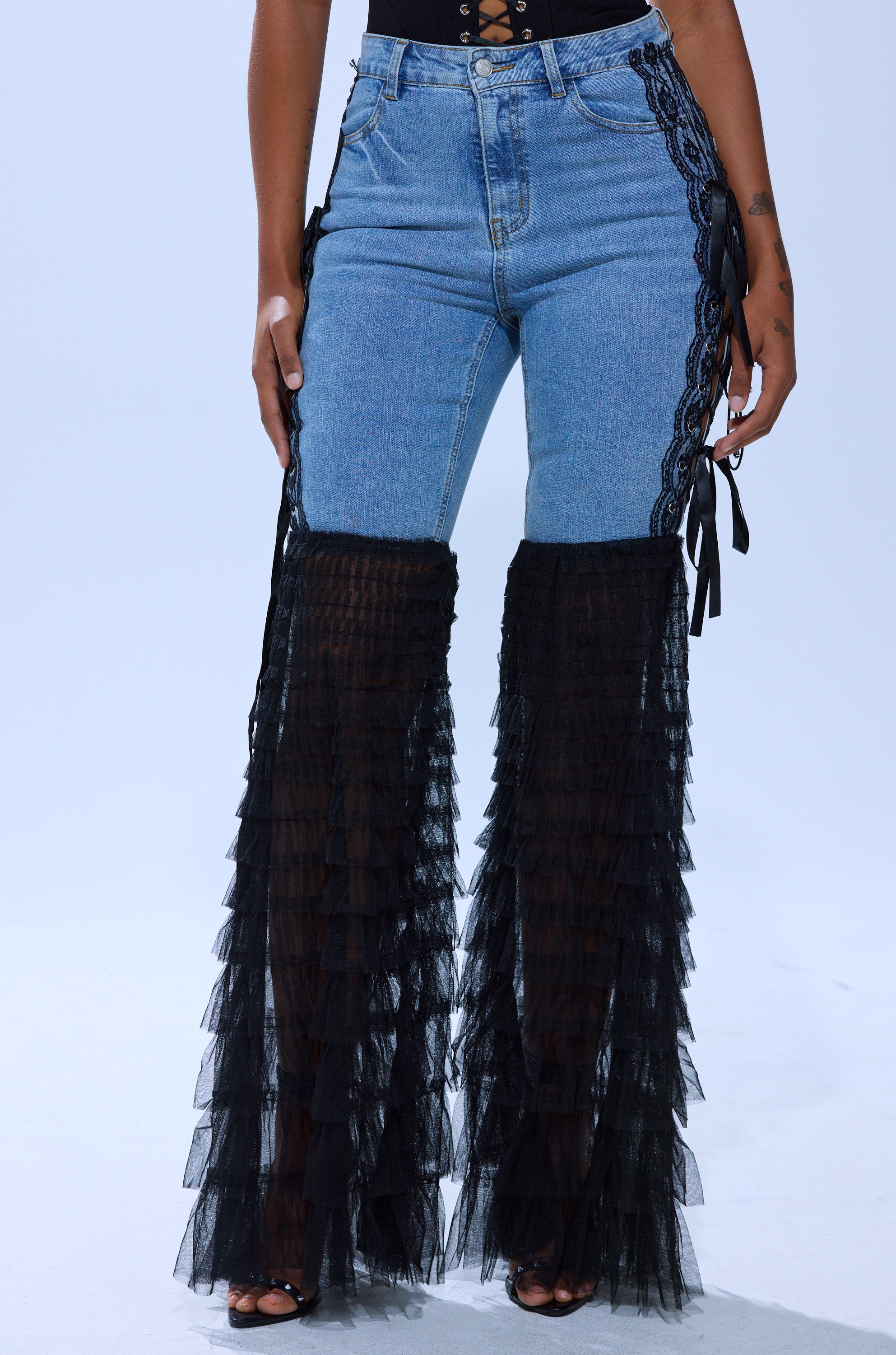 A person wearing CAN'T DECIDE TULLE AND LACE DENIM JEANS, featuring high-waisted ruffled flares, black lace-up sides, and sheer black tulle panels from knee to ankle for a bold, fashion-forward look.